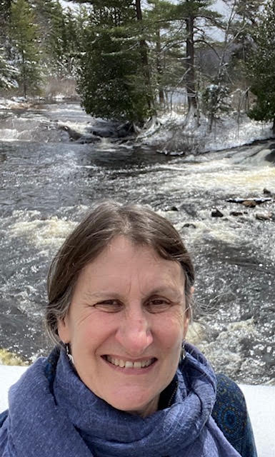 White woman with a purple scarf smiles, outside in front of a rushing river. 