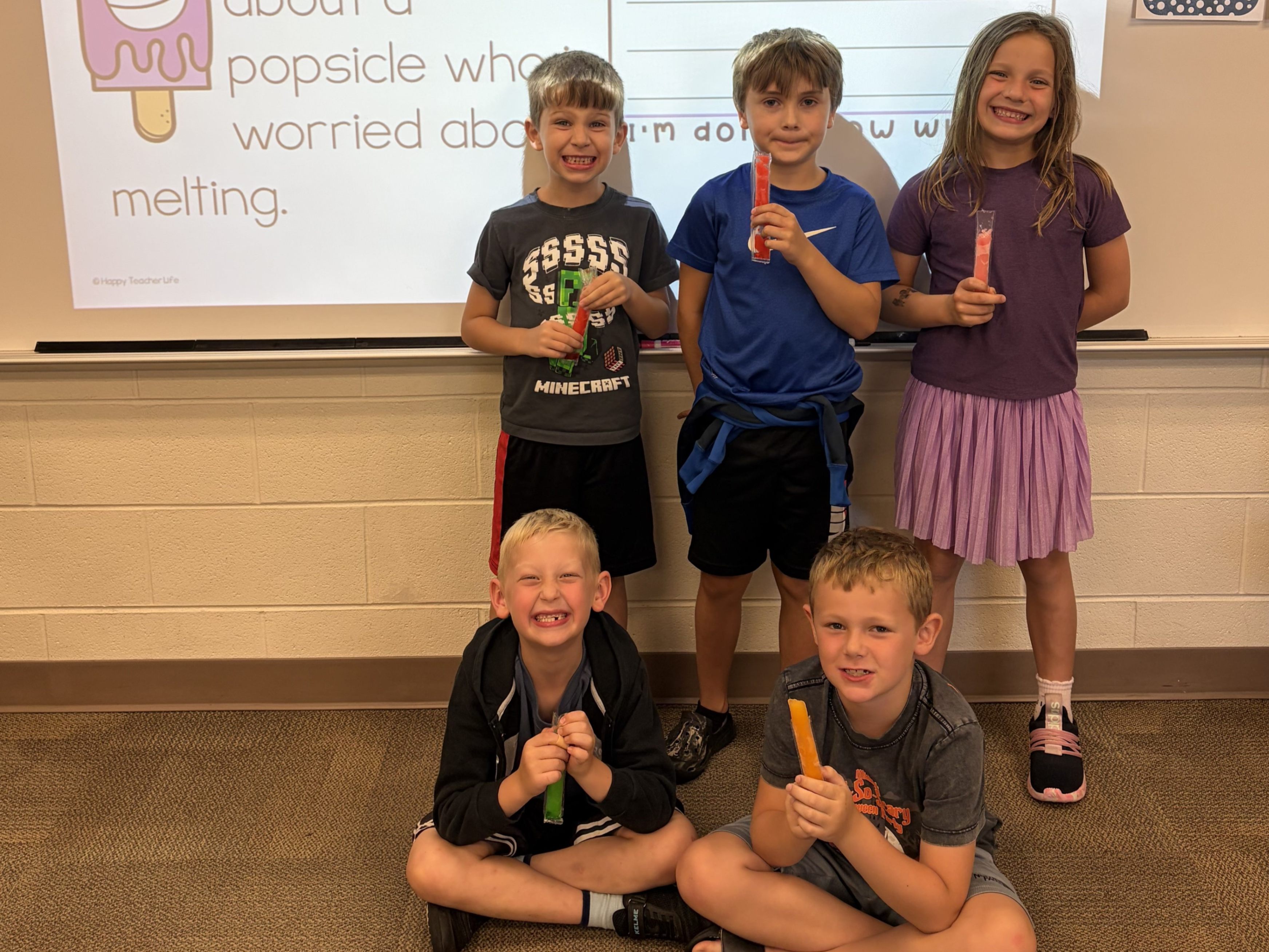 Five young students are holding popsicles in front of a board displaying a writing prompt. Three students are standing and two are sitting on the floor in front of them.