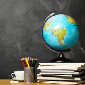 A globe sitting on a stack of books along with a container of colors pencil and a cup and saucer sitting on a desk in front of a blackboard