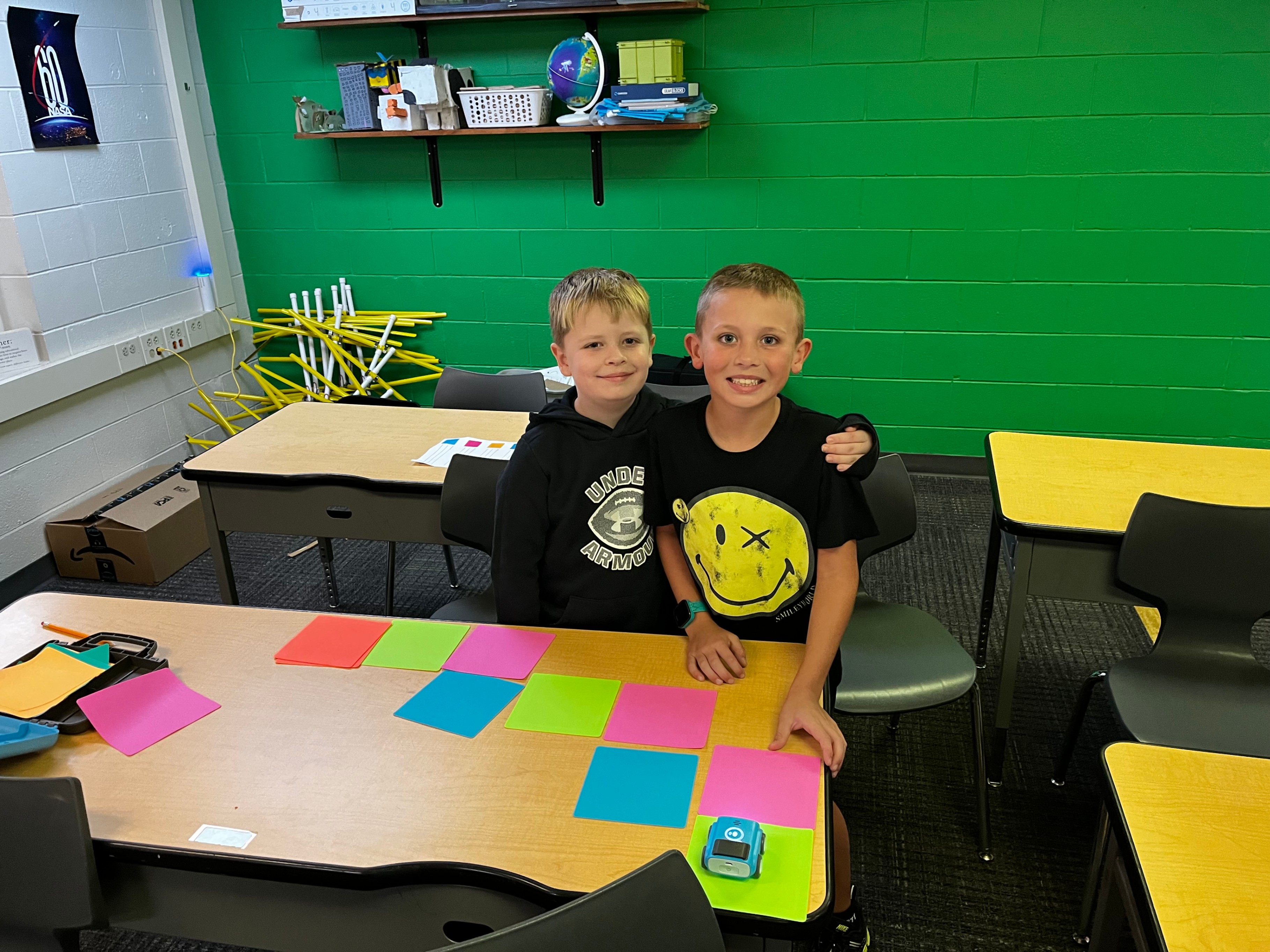 Two student stand with their arms around each other with colored tiles in fron of them