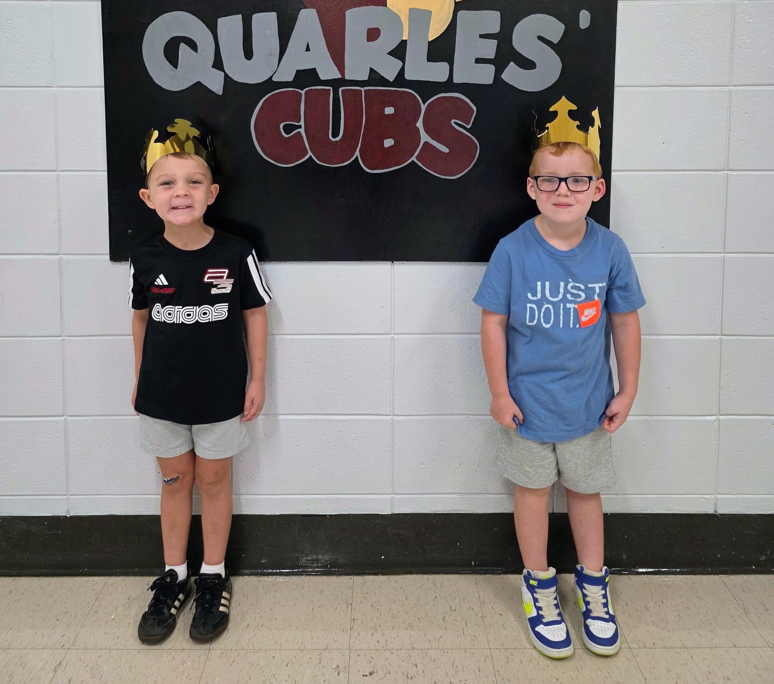 Two elementary‑aged children stand side by side in a school hallway beneath a sign that reads “QUARLES’ CUBS.” Both wear shiny gold paper crowns. The child on the left wears a black Adidas shirt, gray shorts, and black shoes. The child on the right wears a blue Nike “JUST DO IT.” shirt, gray shorts, and blue‑and‑yellow sneakers. They smile in front of a white tiled wall with a black panel displaying the sign, creating a simple, celebratory recognition moment.