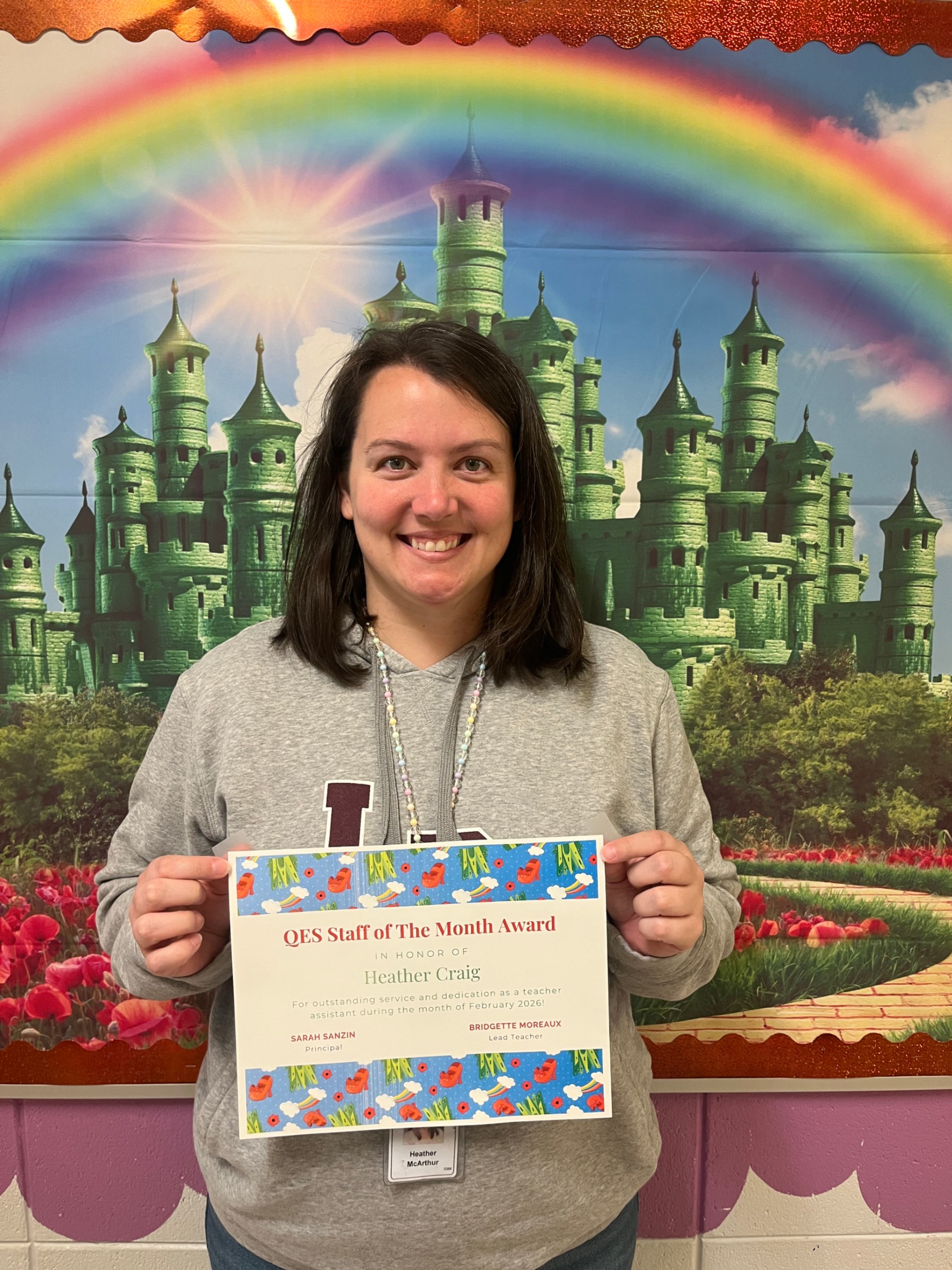 A staff member stands smiling in front of a colorful backdrop featuring a green castle, a rainbow, and a field of red flowers. She holds a certificate recognizing her as QES Staff of the Month, awarded to Heather Craig for her dedication to supporting students and staff. The certificate includes signatures and the date February 2026.