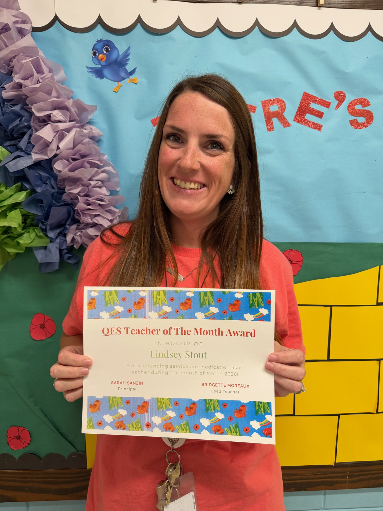 A woman stands smiling in front of a colorful bulletin board while holding a certificate. She wears a coral shirt with a lanyard and keys around her neck. The bulletin board has a blue background, a scalloped white border, a cartoon bluebird, large red letters spelling “TRE’S,” and tissue‑paper flowers in purple, blue, and green. A yellow brick‑pattern border is on the right side. The certificate reads “QES Teacher of the Month Award” and recognizes Lindsey Stout for outstanding service and dedication as a teacher during March 2026, signed by the principal and lead teacher.