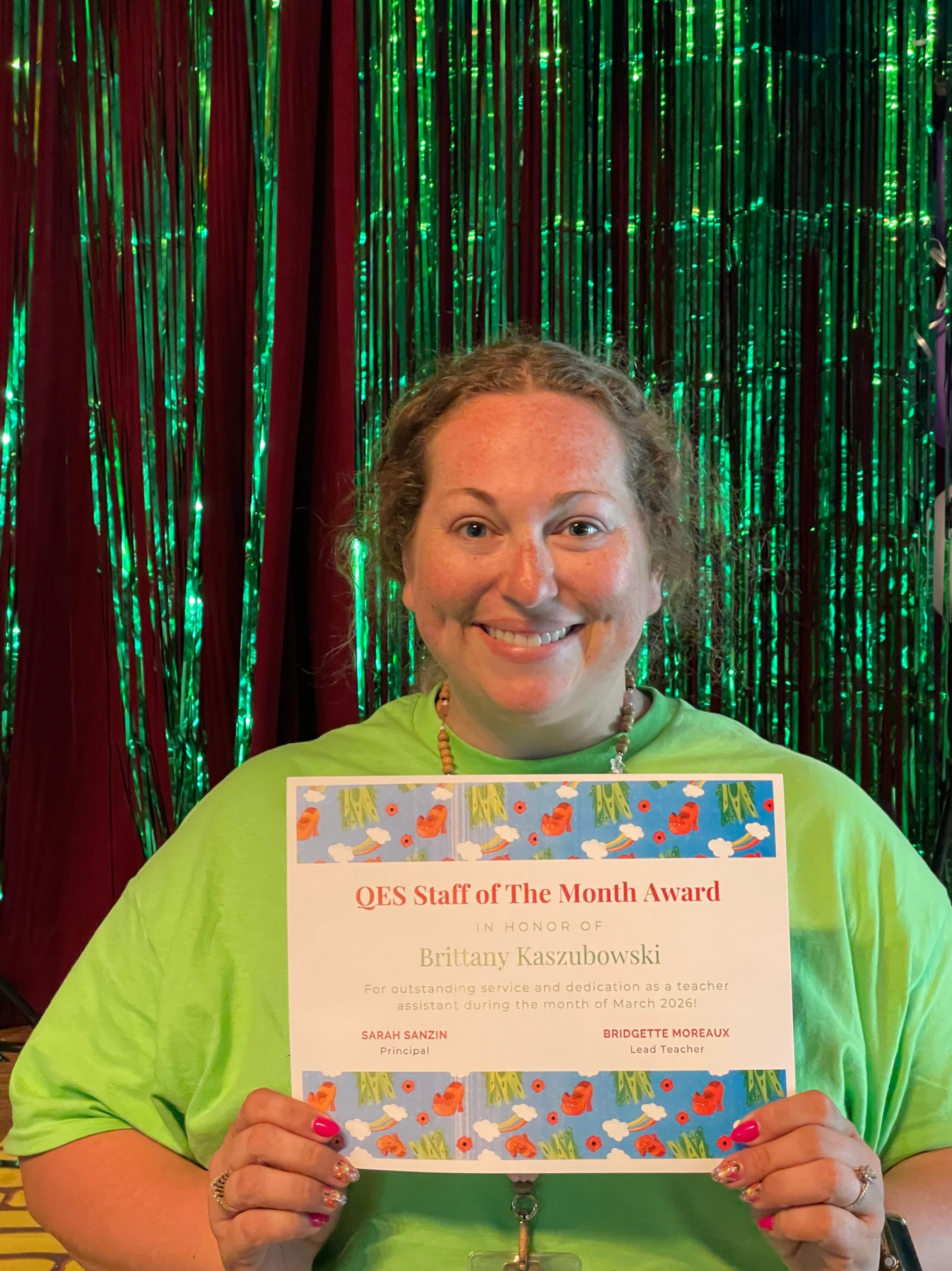    A person stands smiling in front of a backdrop made of shiny green metallic streamers while holding a colorful certificate. They wear a light green shirt. The certificate has a cartoon animal border and reads “QES Staff of the Month Award” honoring Brittany Kaszubowski for outstanding service and dedication as a teacher assistant , signed by the lead teacher and principal.