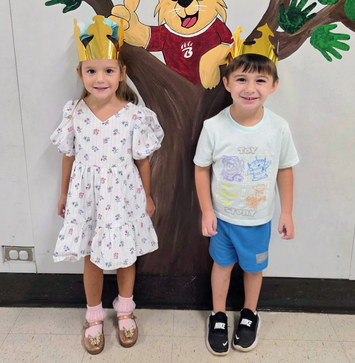 Two young children wearing gold paper crowns stand side by side in front of a colorful wall mural featuring a cartoon animal in a tree. One child wears a white floral dress with gold shoes, and the other wears a light Toy Story T-shirt, blue shorts, and black sneakers.