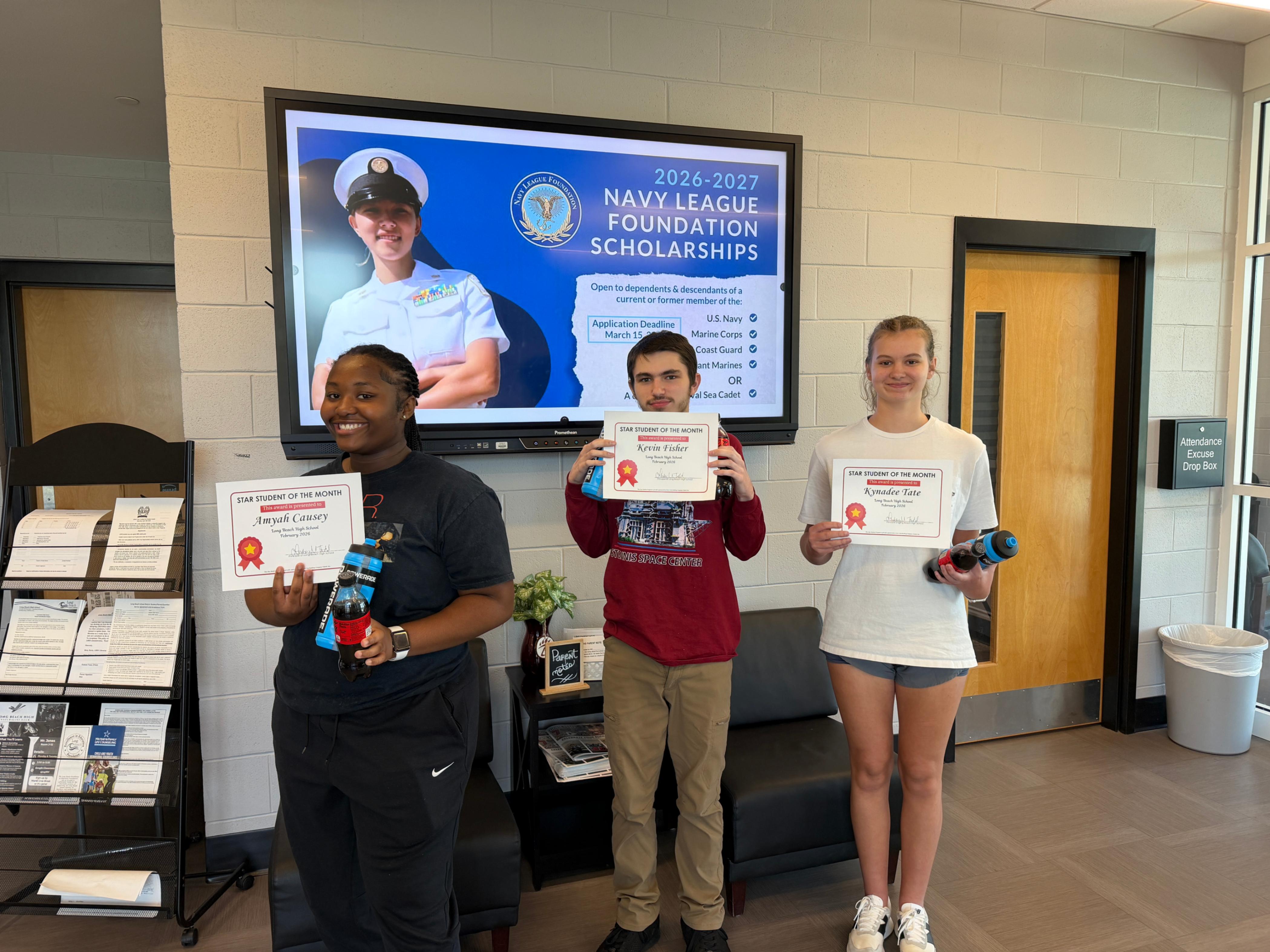 Three students stand indoors holding “Star Student of the Month” certificates and drink bottles. Behind them, a digital display promotes the “2026–2027 Navy League Foundation Scholarships,” listing eligibility for dependents of U.S. Navy, Marine Corps, Coast Guard, Merchant Marines, or Naval Sea Cadet members, with a March 1, 2026 deadline. A wall sign reads “Attendance Excuse Drop Box,” and pamphlets are visible nearby.
