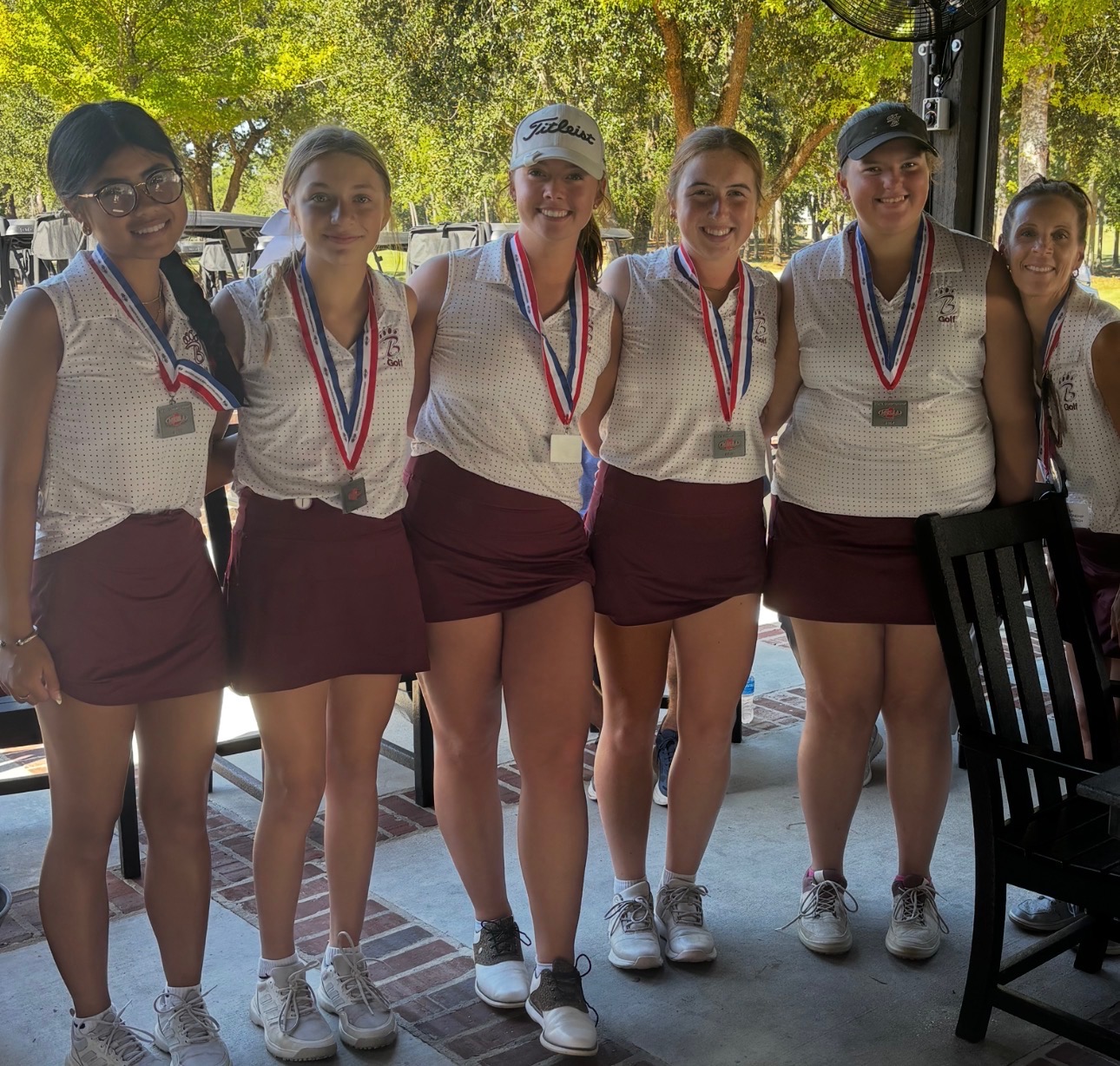 Five student golfers stand together on a golf course wearing bright pink sleeveless shirts, white skirts, and white golf shoes. Three of the golfers wear medals around their necks. The golfer in the center holds a large championship trophy, while another golfer holds a smaller golf-themed trophy. Trees, a wooden fence, and parked school buses are visible in the background on a sunny day.