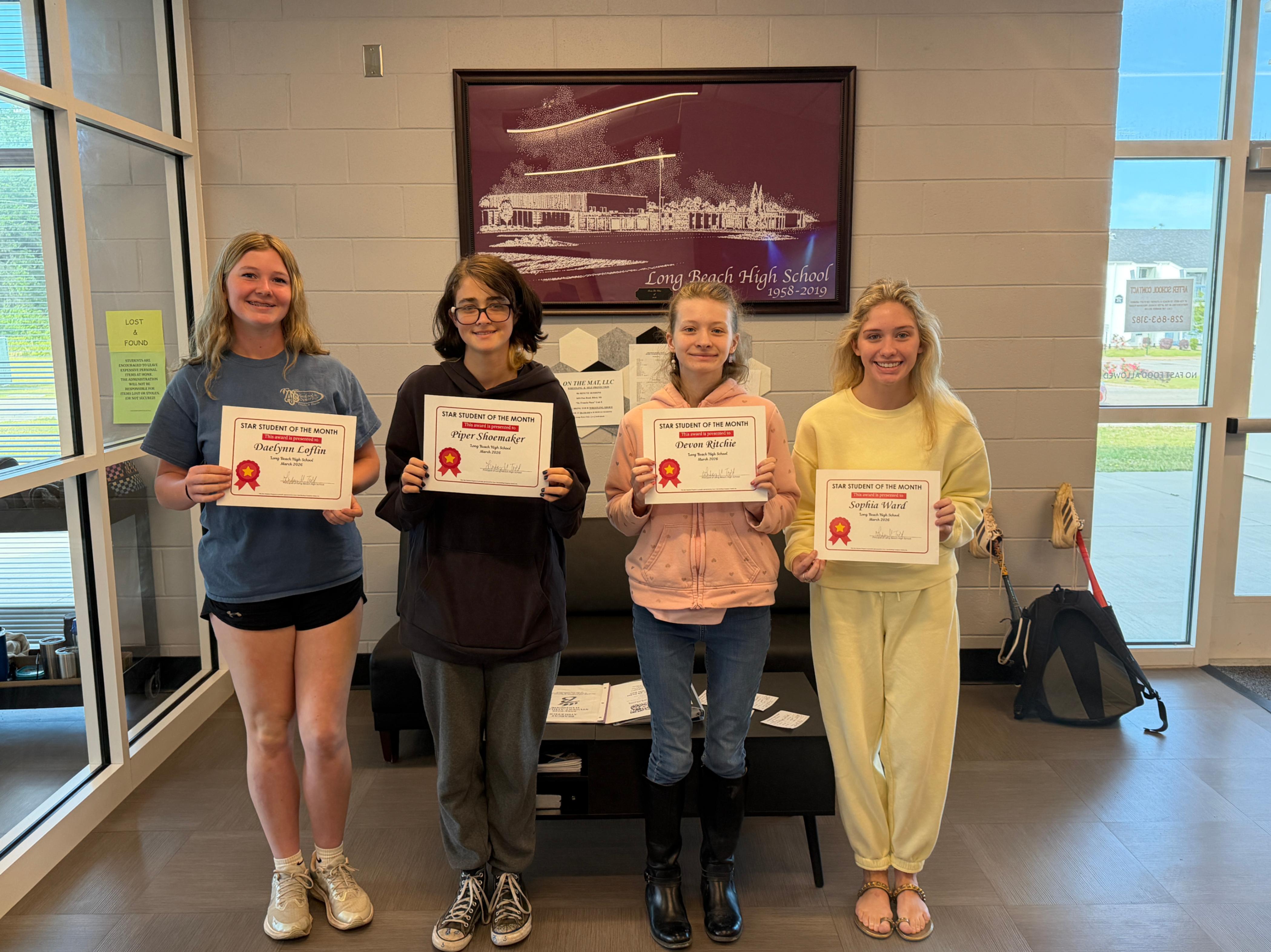 Four students stand indoors holding “Star Student of the Month” certificates. Behind them hangs framed artwork labeled “Long Beach High School 1958–2019.” The students are positioned in a lobby area with a bench, papers on the bench, and a “Lost and Found” sign on the wall. The certificates display the names Danelynn Lafflin, Piper Shoemaker, Oreon Ritchie, and Sophia Wizard.