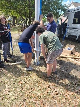 A group of five people work together outdoors to install a tall metal flag pole into the ground. They steady the pole and use tools while standing on a grassy area near a chain-link fence, with trees and a white vehicle in the background.