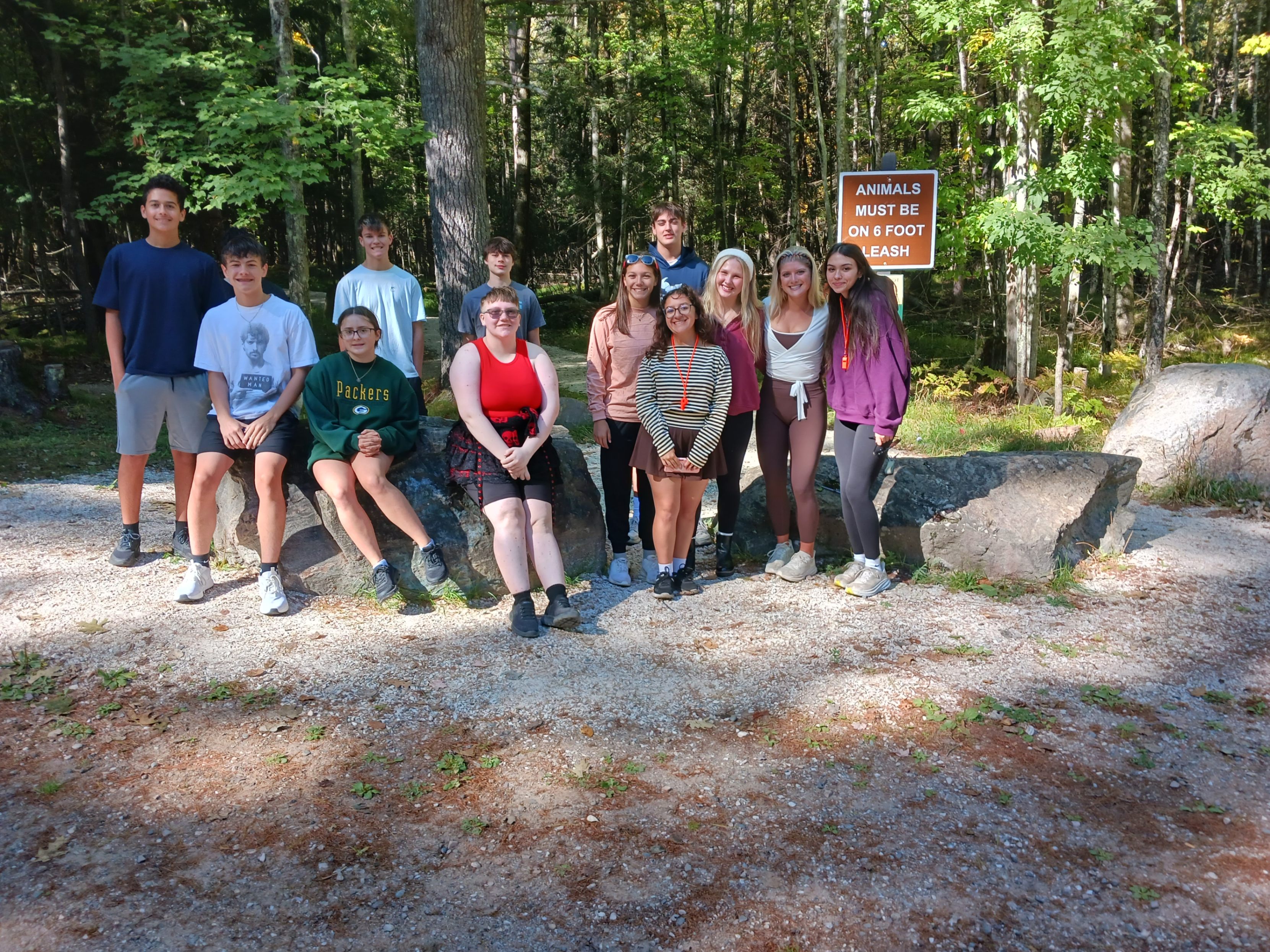 A group of ten high school students posing together outdoors near a large rock formation at the Piers Gorge trailhead. In the background, lush forest trees and a wooden sign read "ANIMALS MUST BE ON 6 FOOT LEASH". Some students are seated on the boulders while others stand beside them, smiling for the camera on a sunny day.