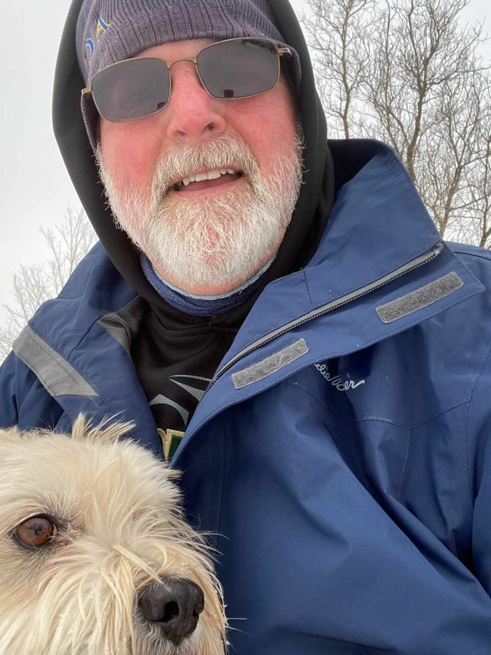 A white man in a winter coat and hat, with a white beard, wearing sunglasses, poses with a white terrier in an outdoor landscape.