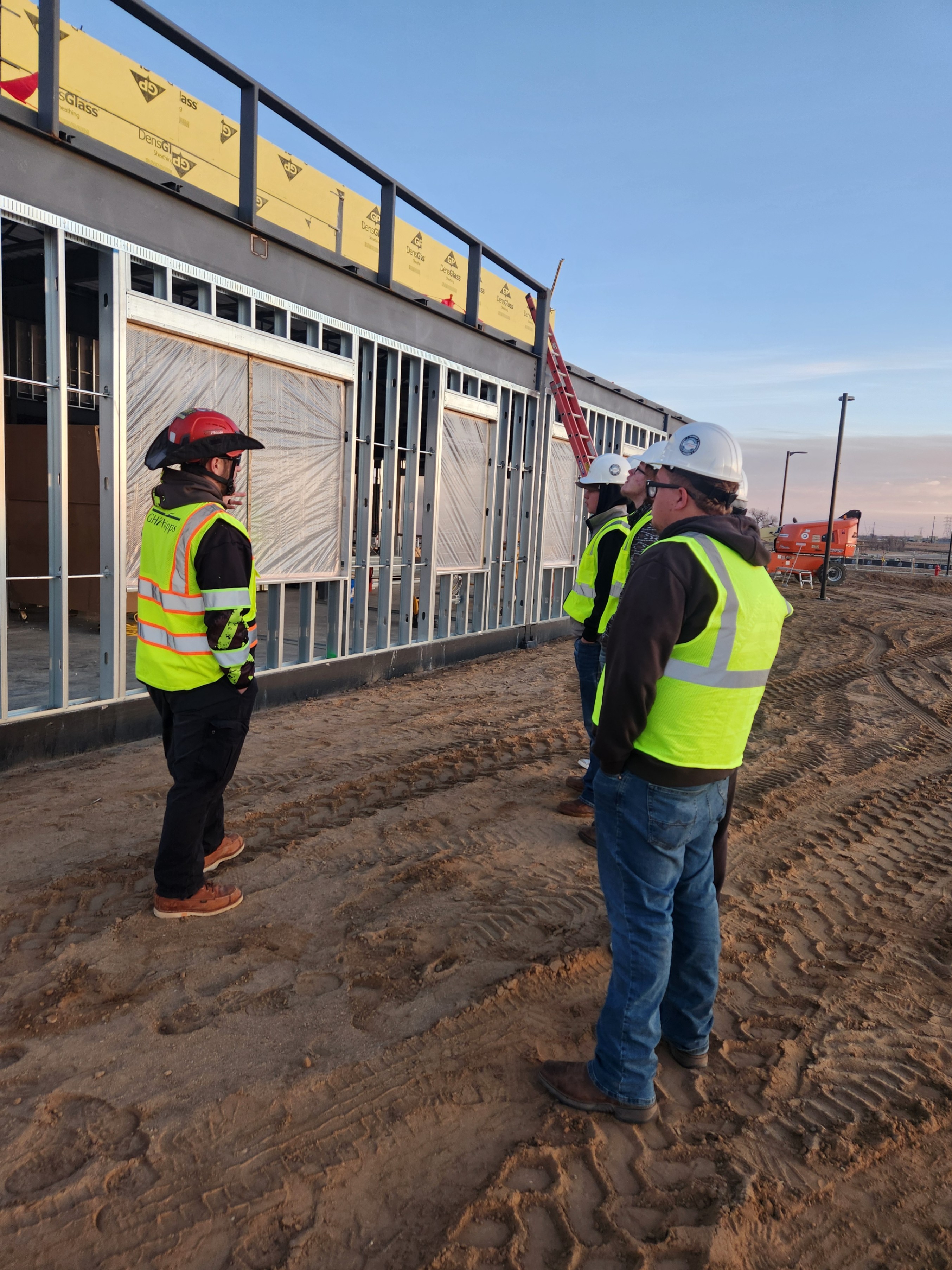 3 male students on a construction site, looking at a building while a team lead is talking to them