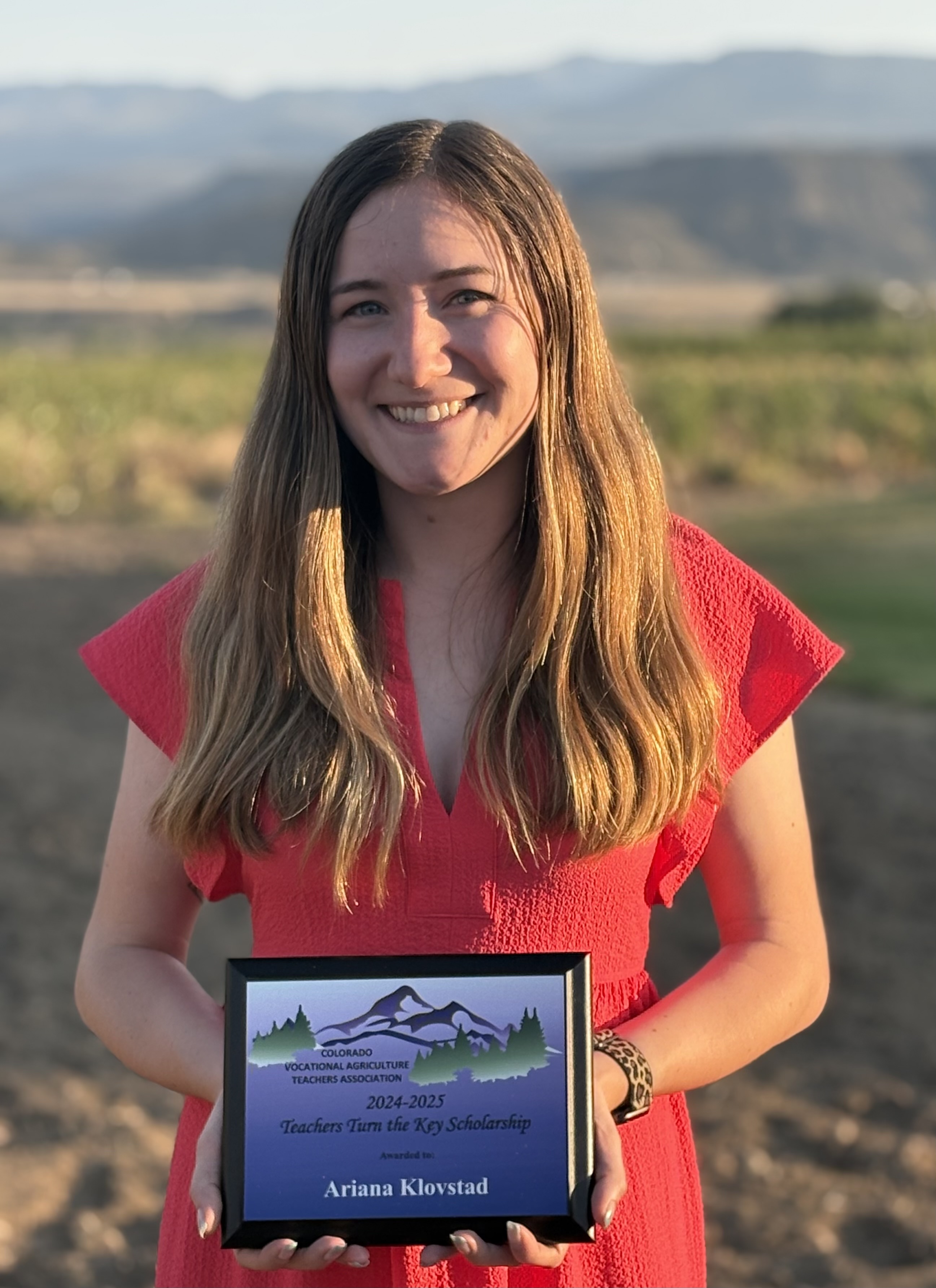 A young woman with long brown hair, wearing a reddish-pink dress, stands outdoors holding a plaque. The plaque reads "Colorado Vocational Agriculture Teachers Association 2024-2025 Teachers Turn the Key Scholarship Awarded to Ariana Klovstad." She is smiling directly at the camera, with a rural landscape of rolling hills and trees visible in the background.