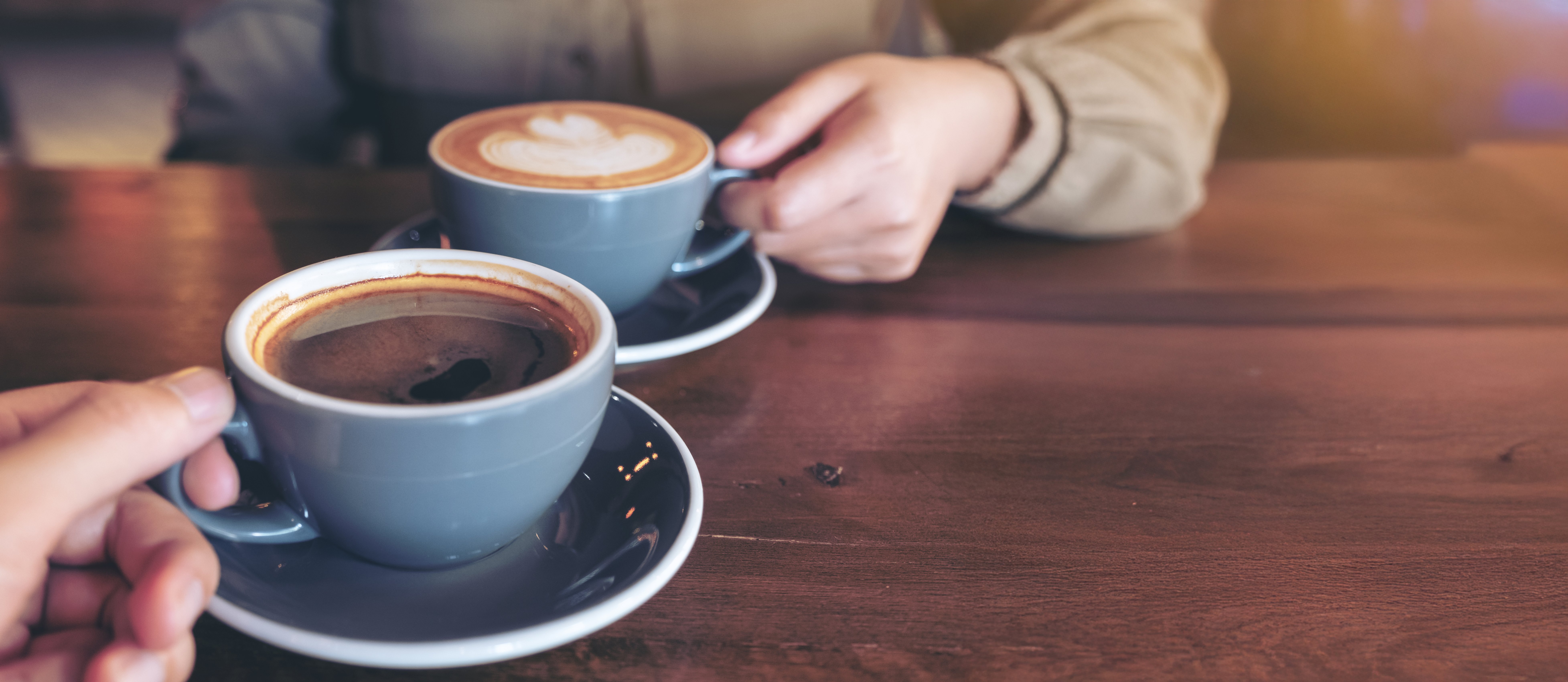 A high-angle, close-up shot of two people holding grey ceramic mugs of coffee with saucers. One person holds a mug of black coffee with a visible reflection, while the other holds a latte with foam art. They are sitting at a wooden table in a dimly lit, cozy cafe.