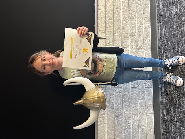 A young girl, holding a Viking helmet and a certificate, stands in front of a brick wall.