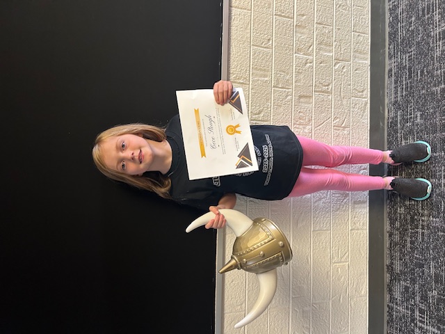 A young girl, holding a Viking helmet and a certificate, stands in front of a brick wall.