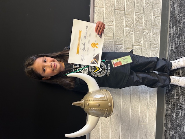 A young girl, holding a Viking helmet and a certificate, stands in front of a brick wall.