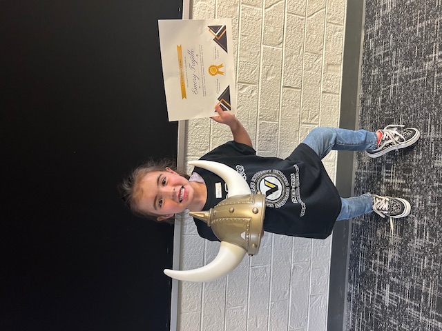 A young girl wearing a black t-shirt, holding a Viking helmet and a certificate, stands in front of a brick wall.