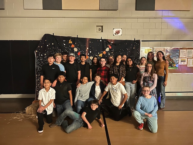 A large group of smiling students posing for a photo in front of a fall-themed backdrop in a gym.