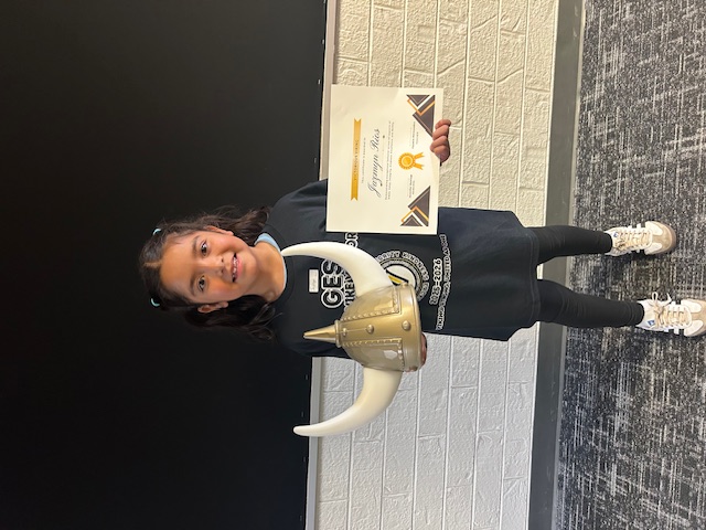 A young girl holding a Viking helmet and a certificate, stands in front of a brick wall.