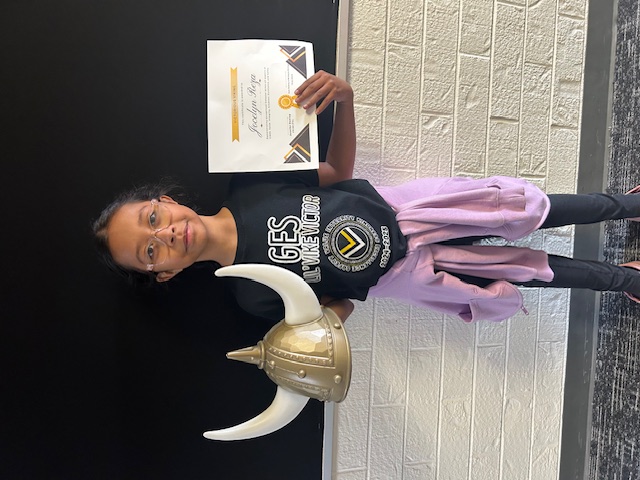 A young girl, holding a Viking helmet and a certificate, stands in front of a brick wall.