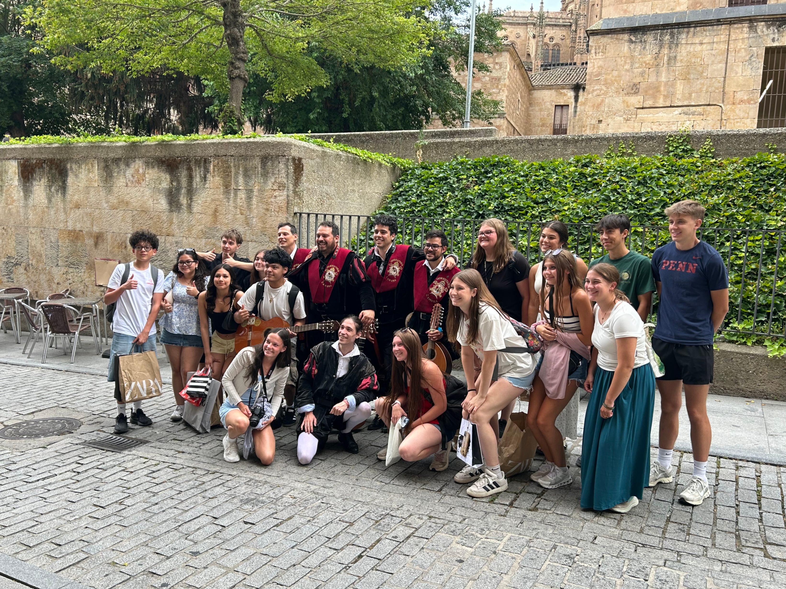 A group of students posing happily on a cobblestone street with members of a traditional Spanish university "Tuna" musical group.