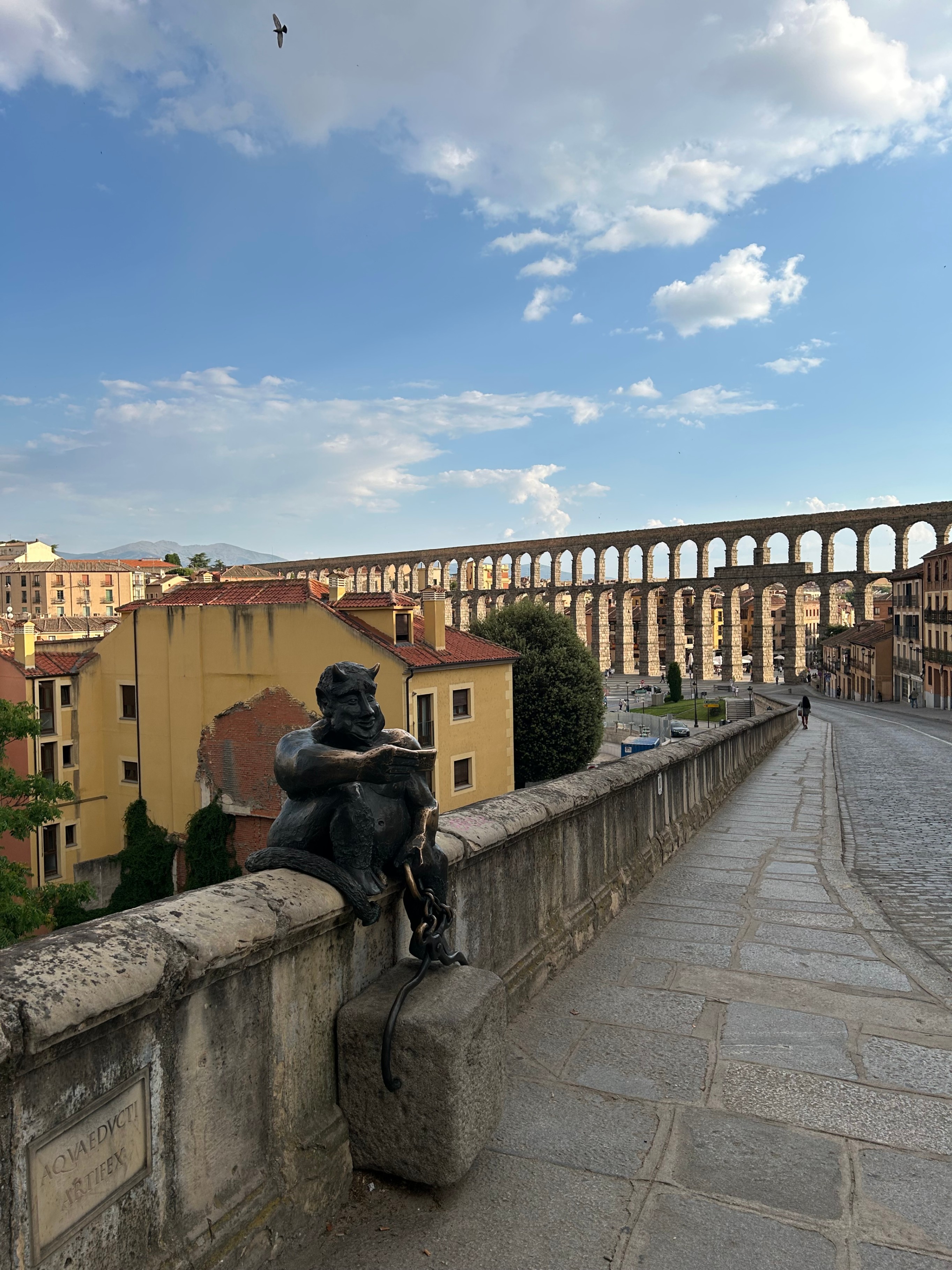 Bronze statue of a smiling devil sitting on a stone wall overlooking the historic Roman Aqueduct of Segovia under a bright sky.