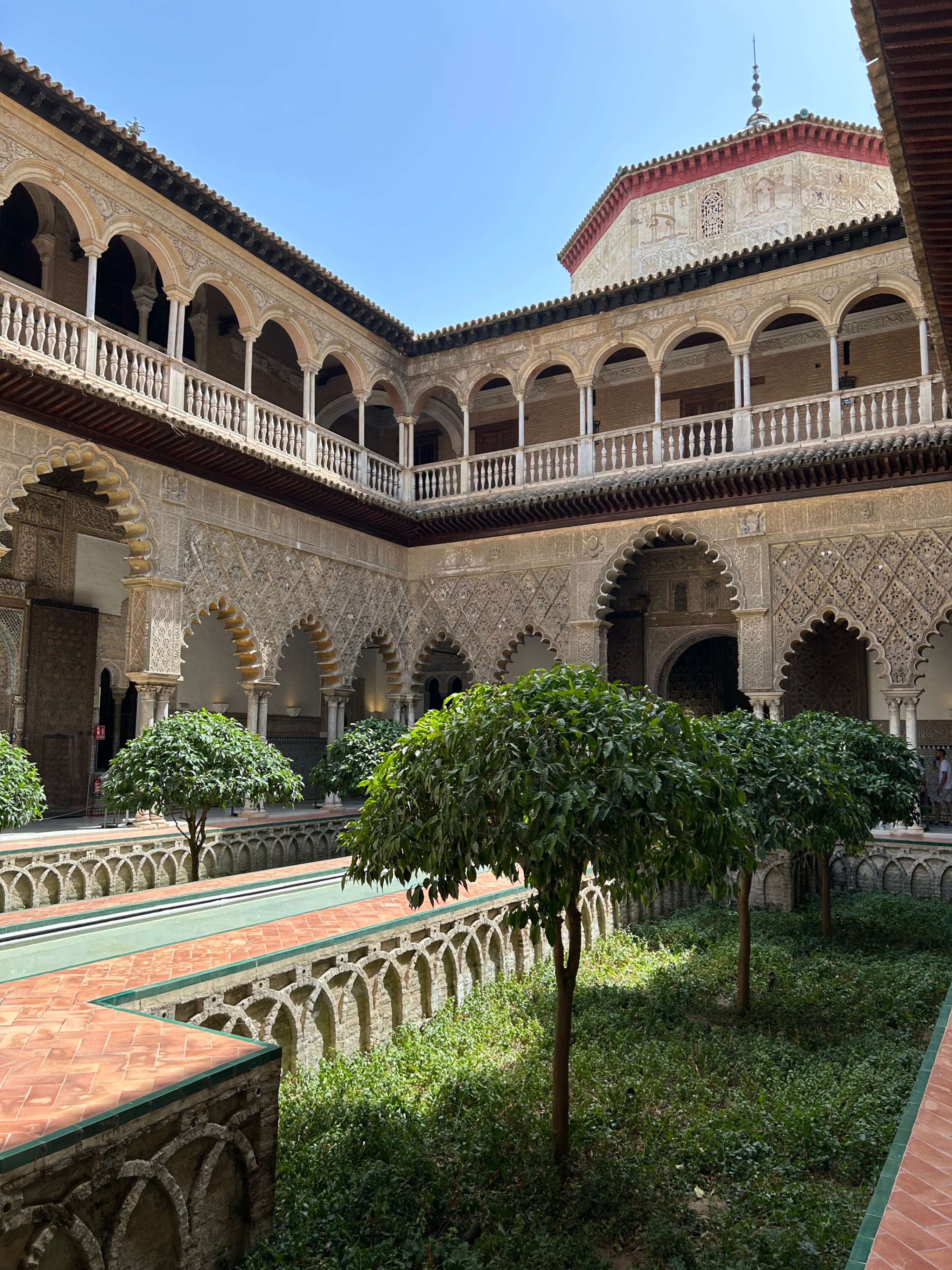 A beautiful interior courtyard featuring ornate Islamic-style arches, a reflecting pool, and lush green trees at the Royal Alcázar of Seville.