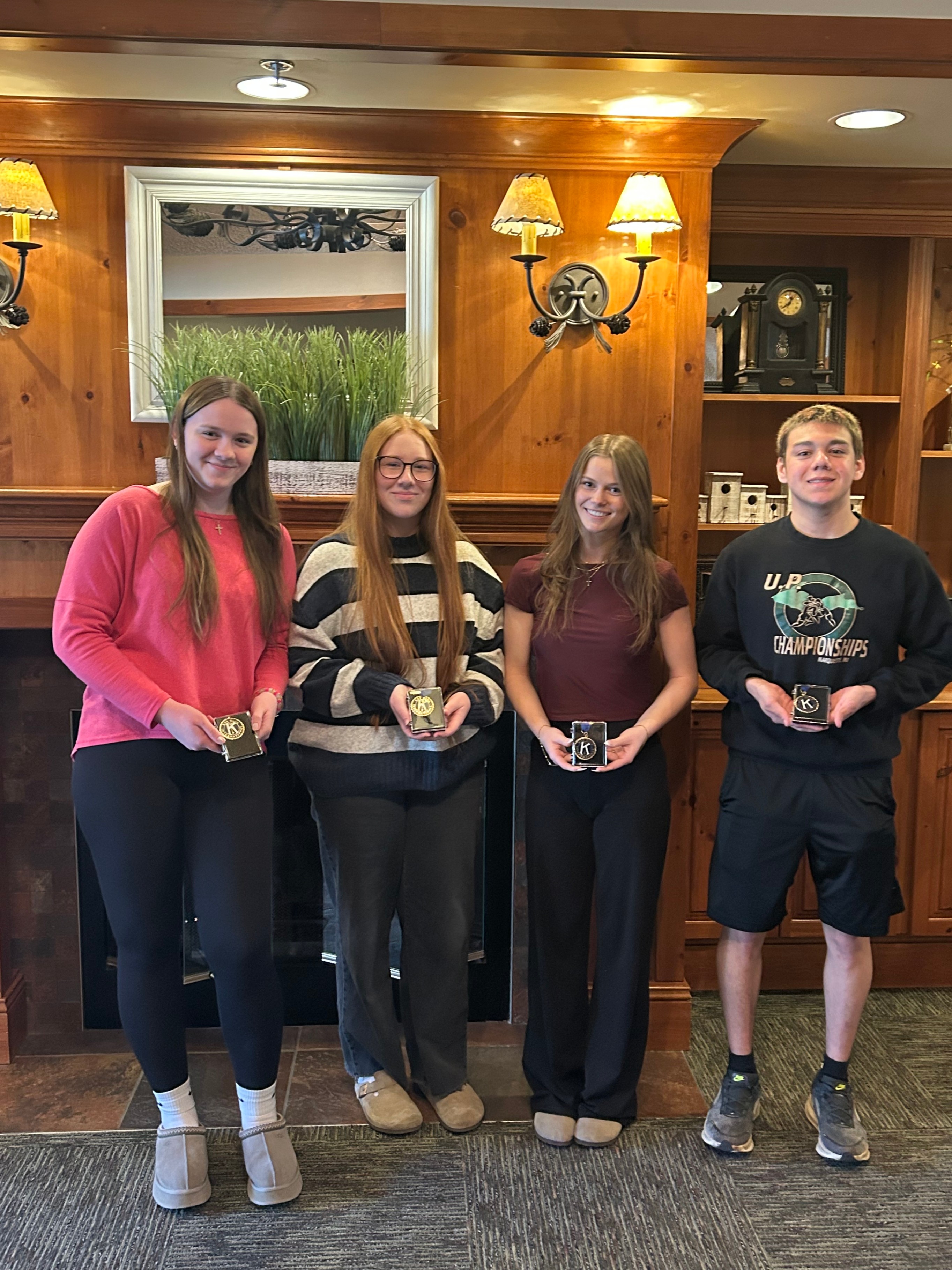 A group of four high school students—three young women and one young man—stand side-by-side in a wood-paneled room in front of a brick fireplace. They are smiling and proudly displaying matching small black award boxes featuring a circular gold "K" emblem. The student on the far right wears a black sweatshirt that reads "U.P. Championships."
