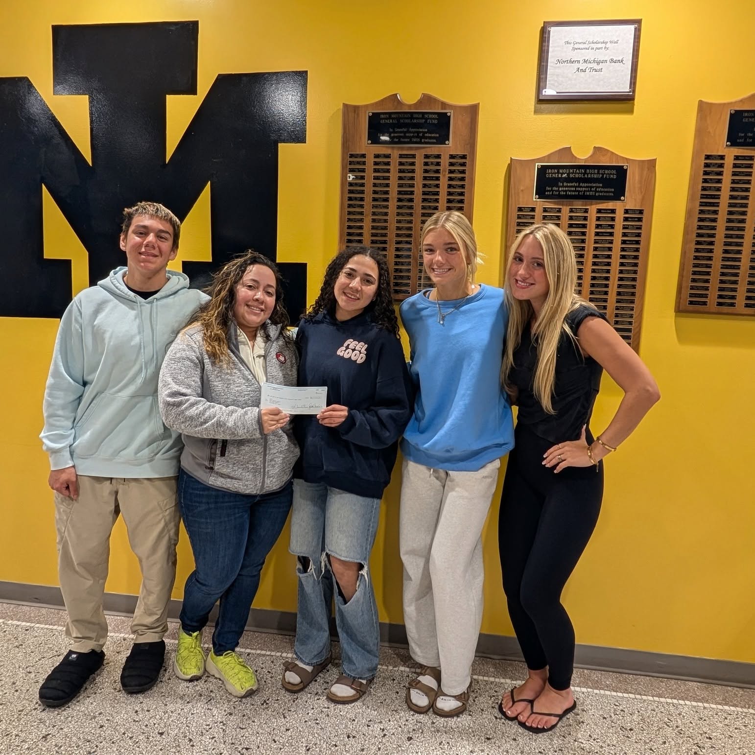 Pictured here Iris Torres- Pagain and Geraldine are accepting a check from Di’Angelo Saldana, Margot McRoberts and Avea Brown posing with a Iris Torres-Pagain and Geraldine Torres holding a donation check in front of a yellow wall with a large black "IM" logo and scholarship plaques.