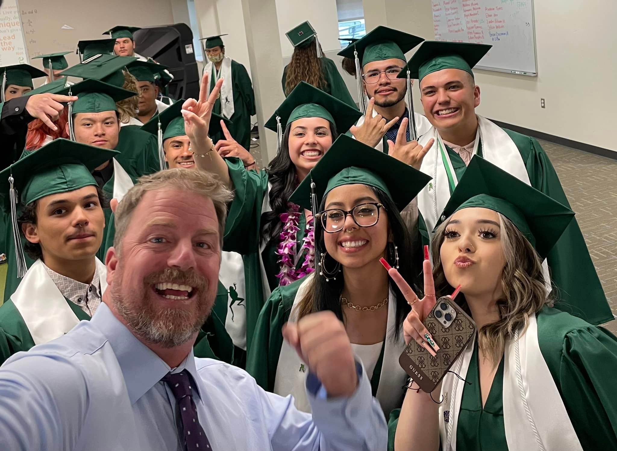 Students posing with the principal for a selfie as they stage in a hallway before graduation ceremony