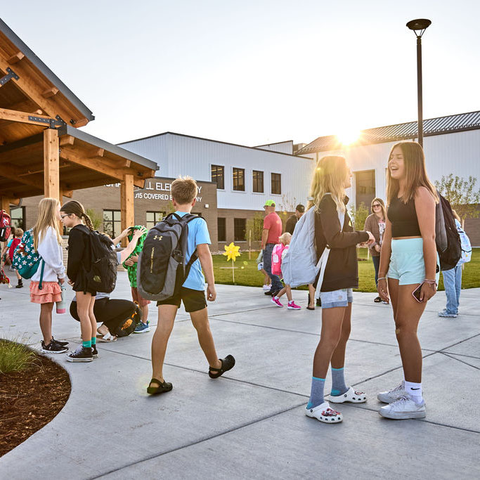Students with backpacks gather outside an elementary school building at sunset.