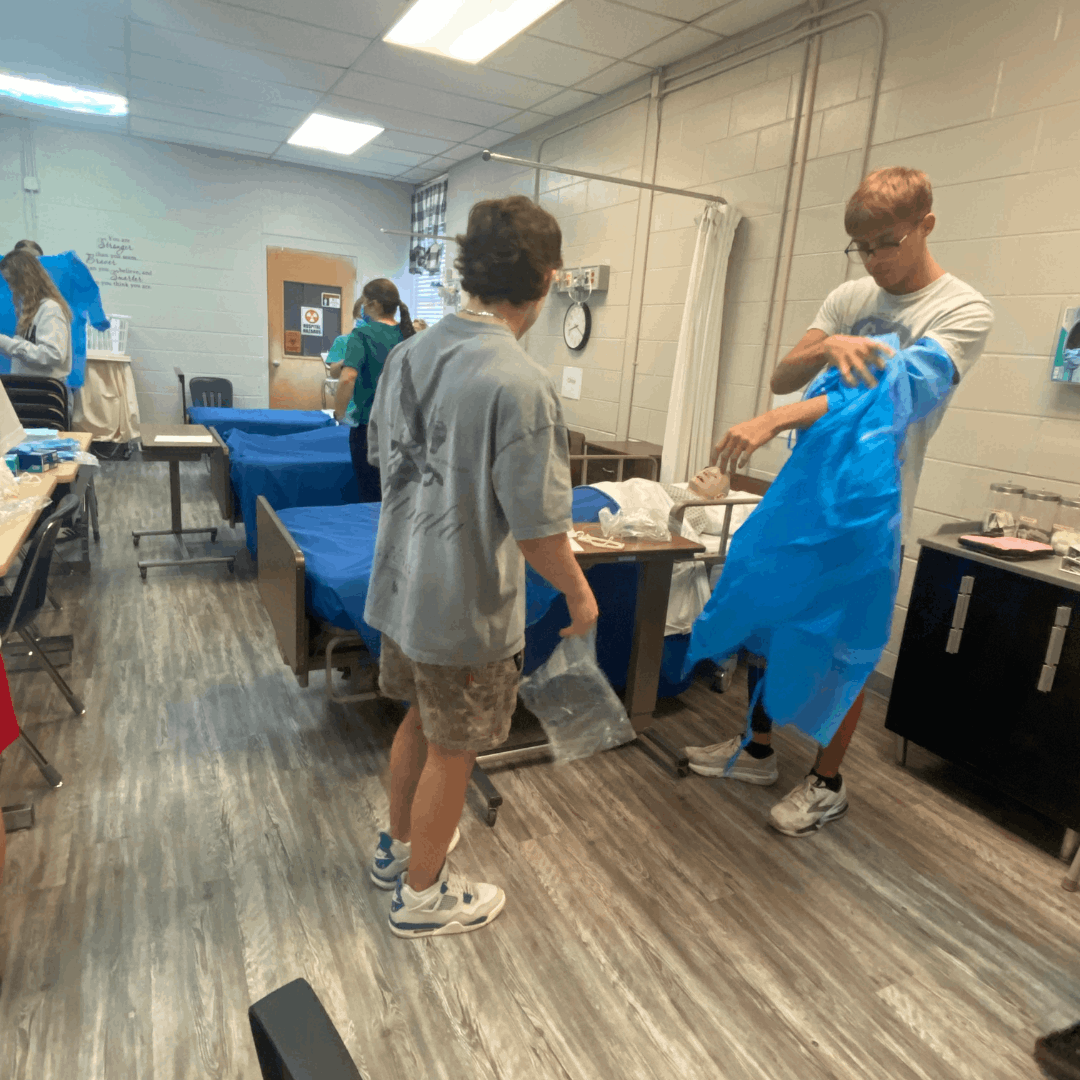 male student donning PPE while another student assists