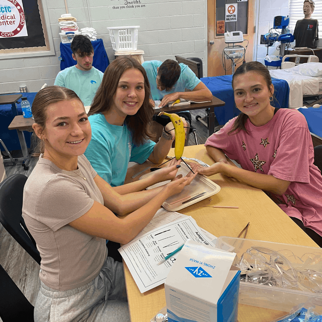 3 female students sitting at a desk, one holding a banana for dissection
