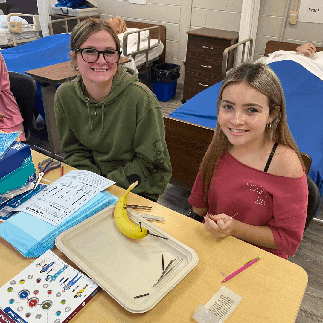 2 female students sitting at a desk, one holding a banana for dissection