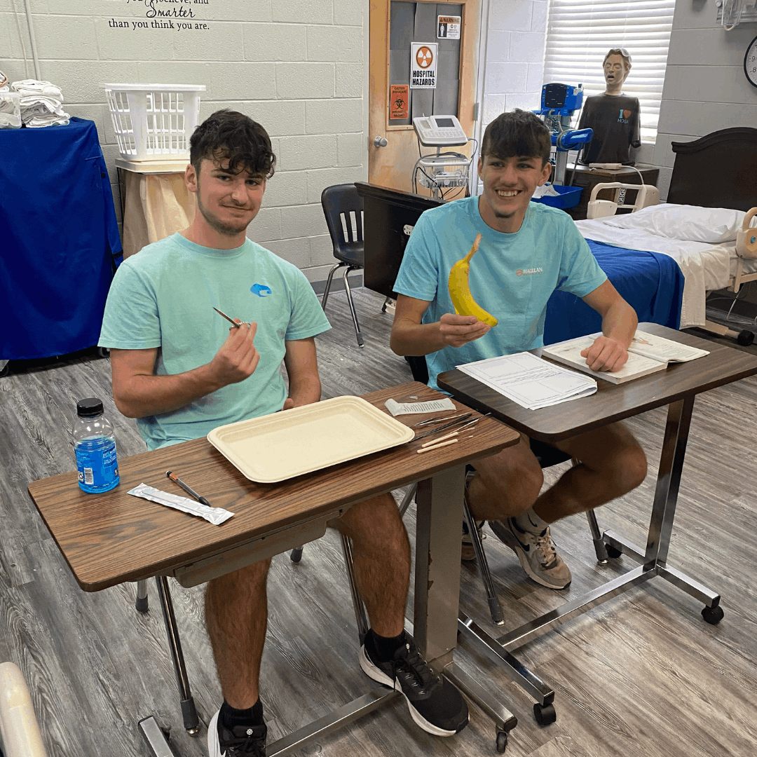 2 male students sitting at desks holding a banana up for practice dissection
