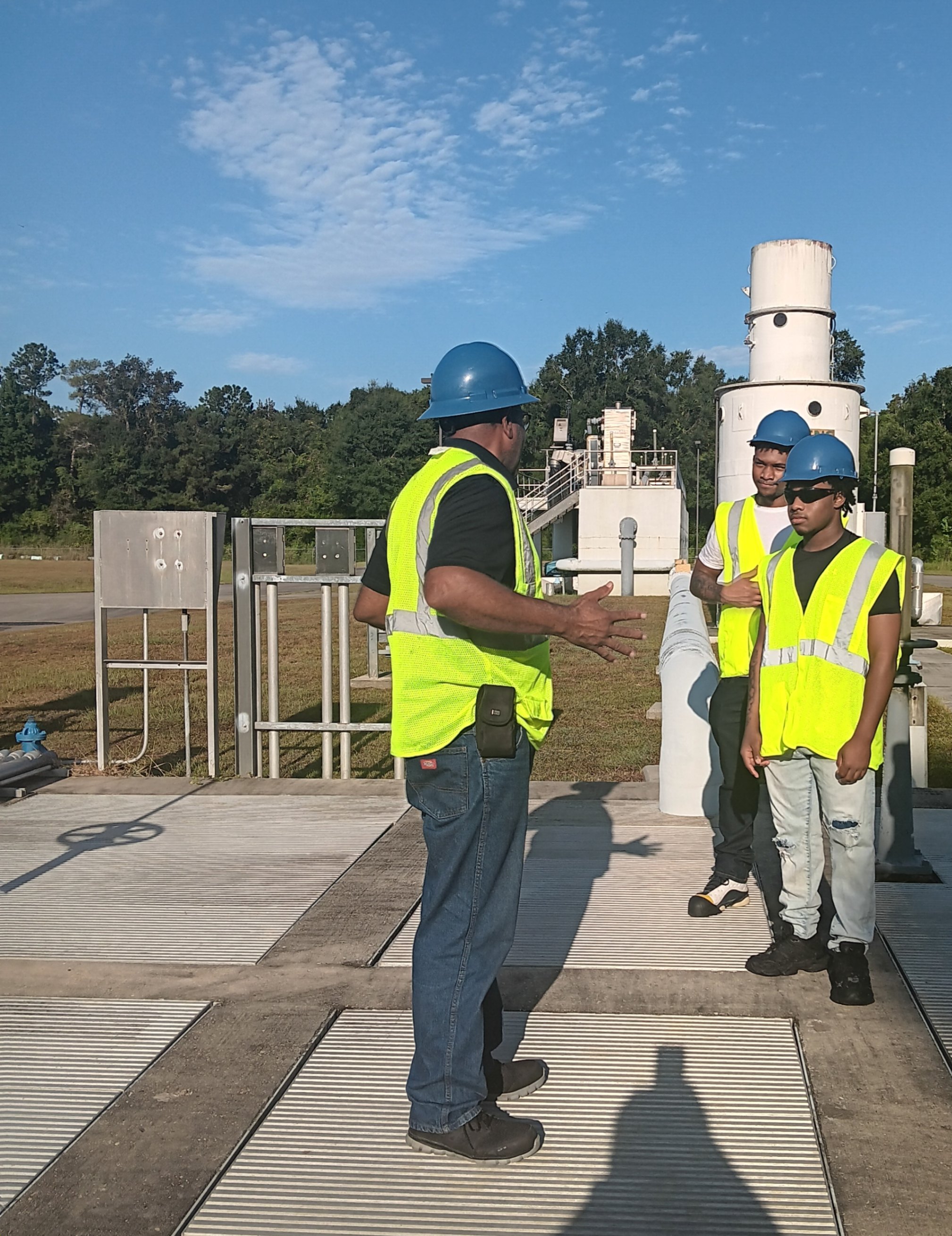 Three workers wearing blue hard hats and bright yellow safety vests stand together at an outdoor industrial facility. One person gestures with both hands while speaking, and the other two listen closely. They stand on metal grating surrounded by pipes and large cylindrical equipment, with trees visible in the background under bright sunlight.