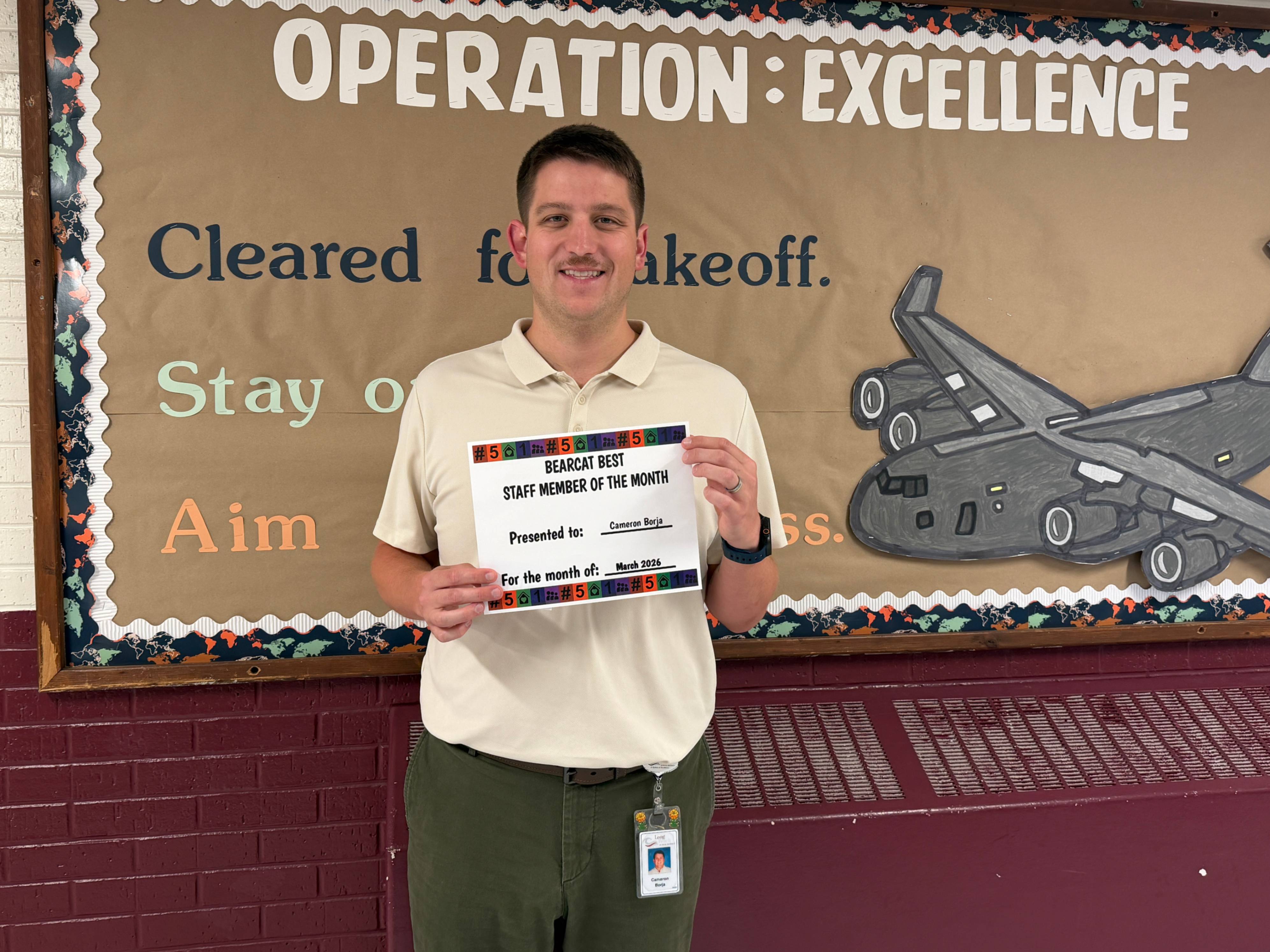 A staff member stands in front of a decorated bulletin board titled “Operation: Excellence,” featuring an airplane graphic and motivational phrases such as “Cleared for takeoff,” “Stay on course,” and “Aim for success.” The person holds a certificate that reads “Bearcat Best Staff Member of the Month,” presented to Cameron Borja for March 2026. The display highlights staff recognition and celebrates excellence.