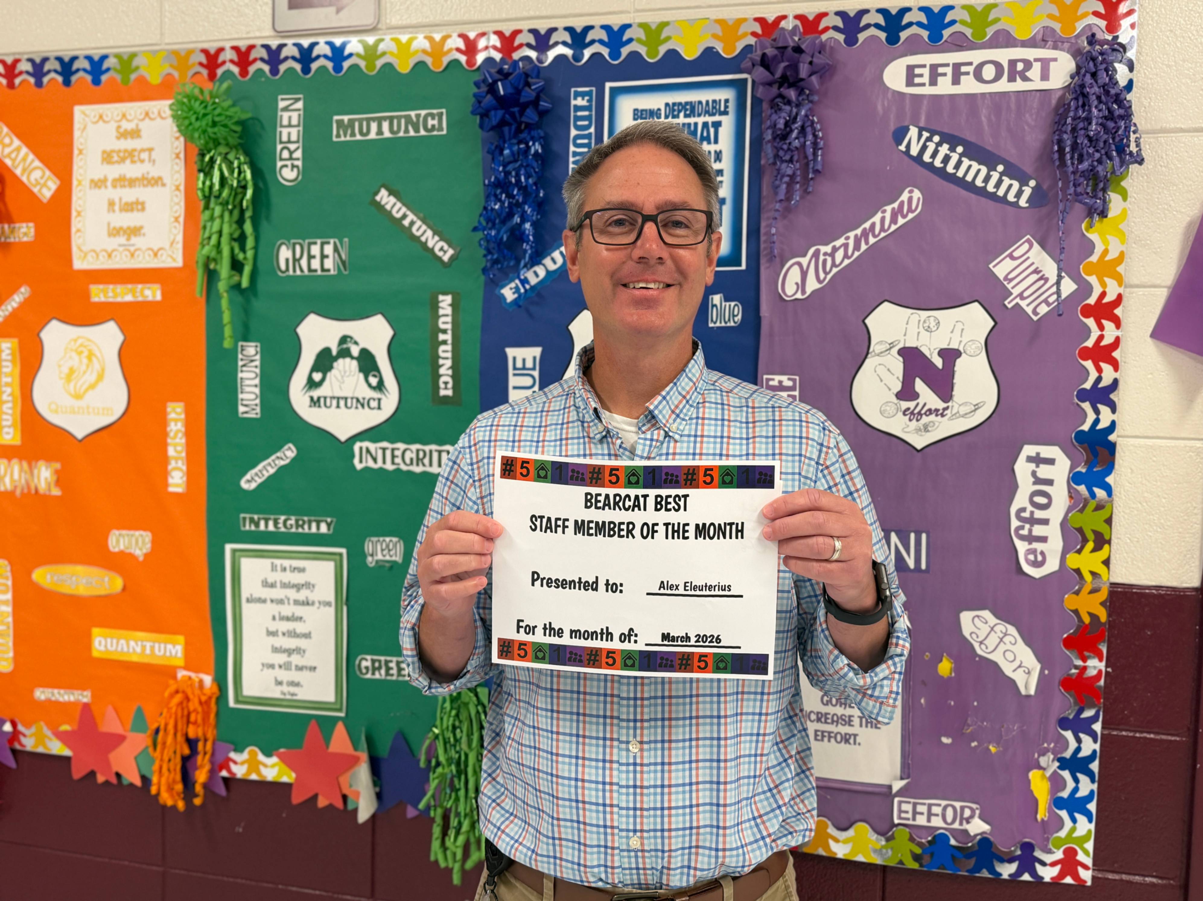 A staff member stands in front of a colorful bulletin board decorated with house names, values, and shield emblems in orange, green, and purple sections. The board includes words such as “Effort,” “Integrity,” “Green,” “Mutunci,” “Nitimini,” “Quantum,” and “Orange,” along with tassel decorations. The person holds a certificate titled “Bearcat Best Staff Member of the Month,” presented to Alex Eleuterius for March 2026. The scene highlights staff recognition and celebrates excellence.