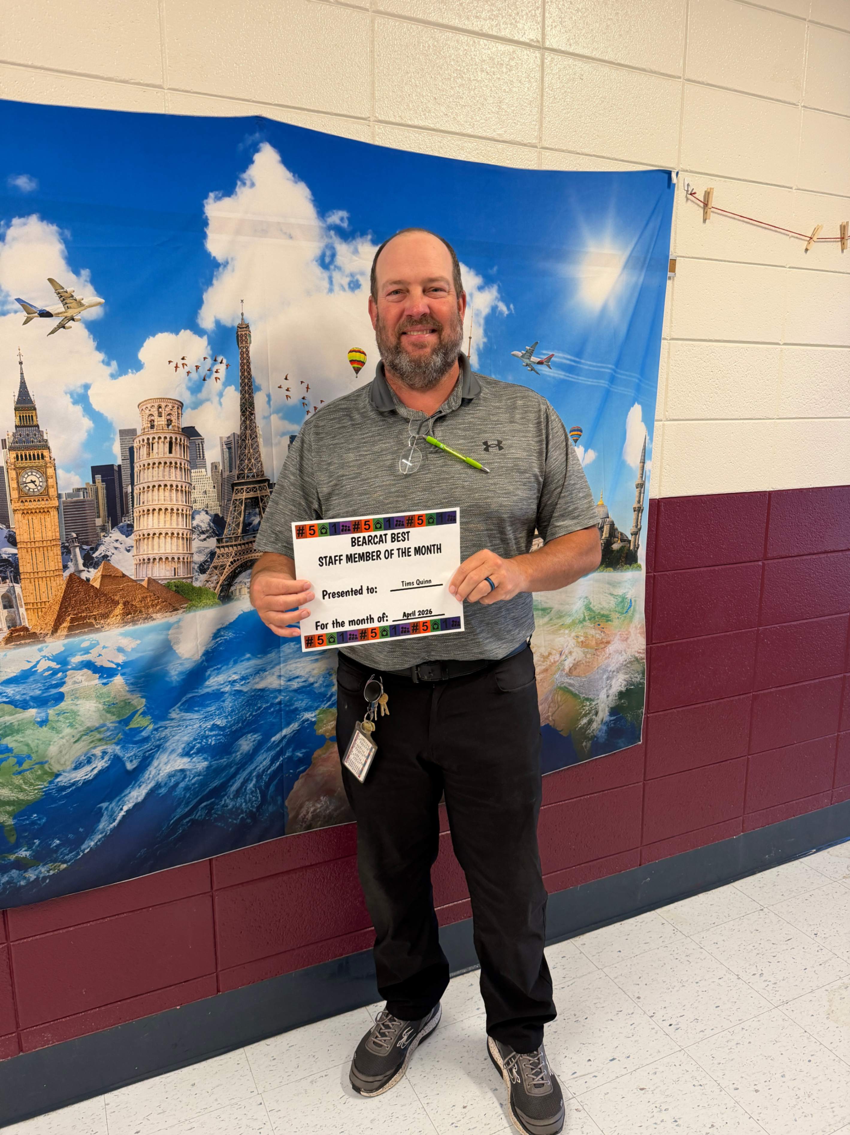 An adult stands indoors in front of a colorful backdrop featuring illustrations of global landmarks, airplanes, and a view of Earth from space. They smile while holding a certificate titled “Staff Member of the Month,” presented to  for the month Tims Quinn of March. The scene conveys recognition and celebration in a school or workplace setting.