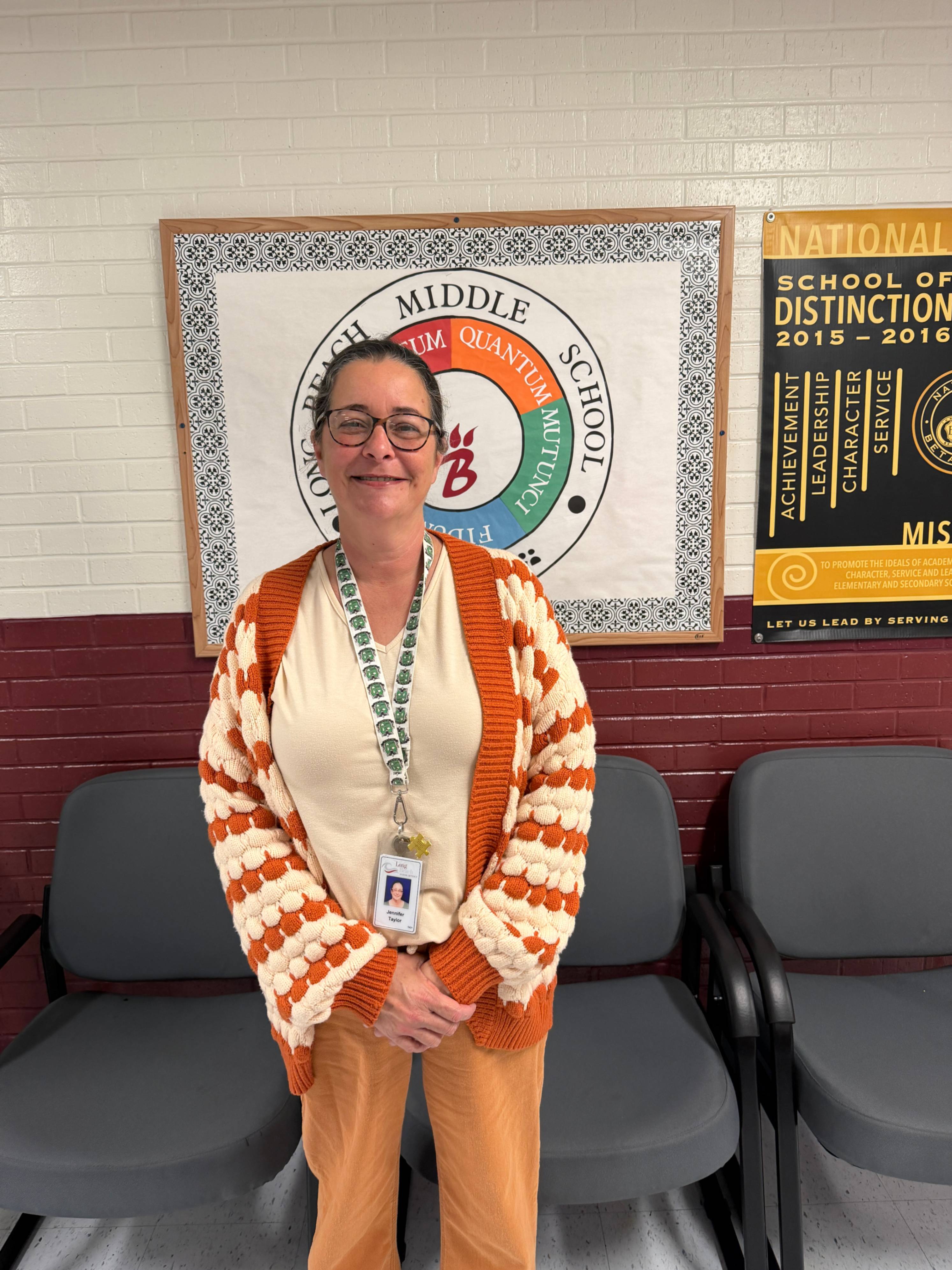 An individual stands indoors in front of a wall displaying school signage. They wear glasses, a textured orange‑and‑cream cardigan over a cream shirt, tan pants, and a lanyard with an ID badge. Behind them, a framed sign reads “Long Beach  Middle School” with a circular emblem containing the words “Quantum Mutunci” and “Fidums.” To the left, a gold and black plaque recognizes the school as a “National School of Distinction 2015–2016” and highlights mission, service, character, leadership, and achievement. Several gray chairs line the wall.