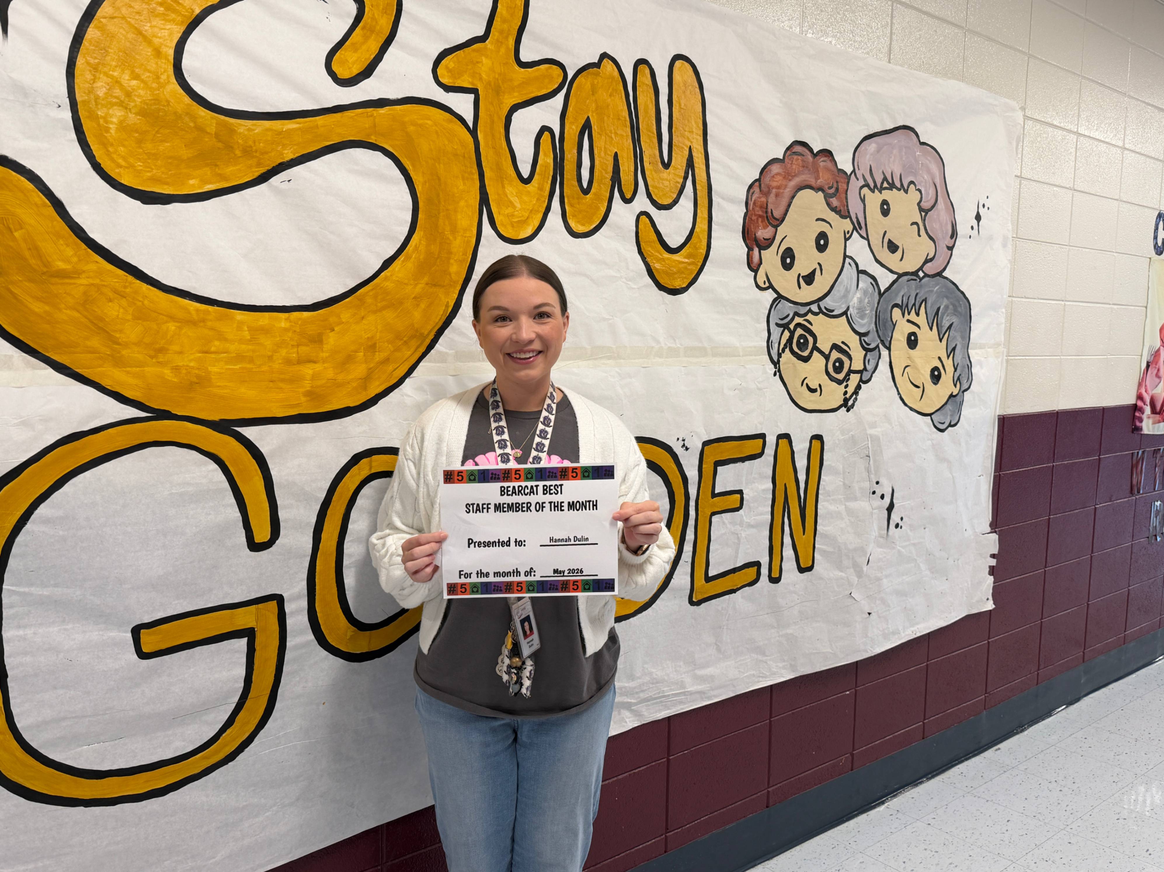 A person stands in a school hallway in front of a large hand-painted banner that reads “Stay Golden” in bold yellow letters, with cartoon drawings of children’s faces on the right side. The person is holding a certificate that reads “Bearcat Best Staff Member of the Month,” with the name “Hannah Dulin” and “May 2026” visible. The hallway has tiled floors and a maroon lower wall, and the person is wearing a lanyard with an ID badge.