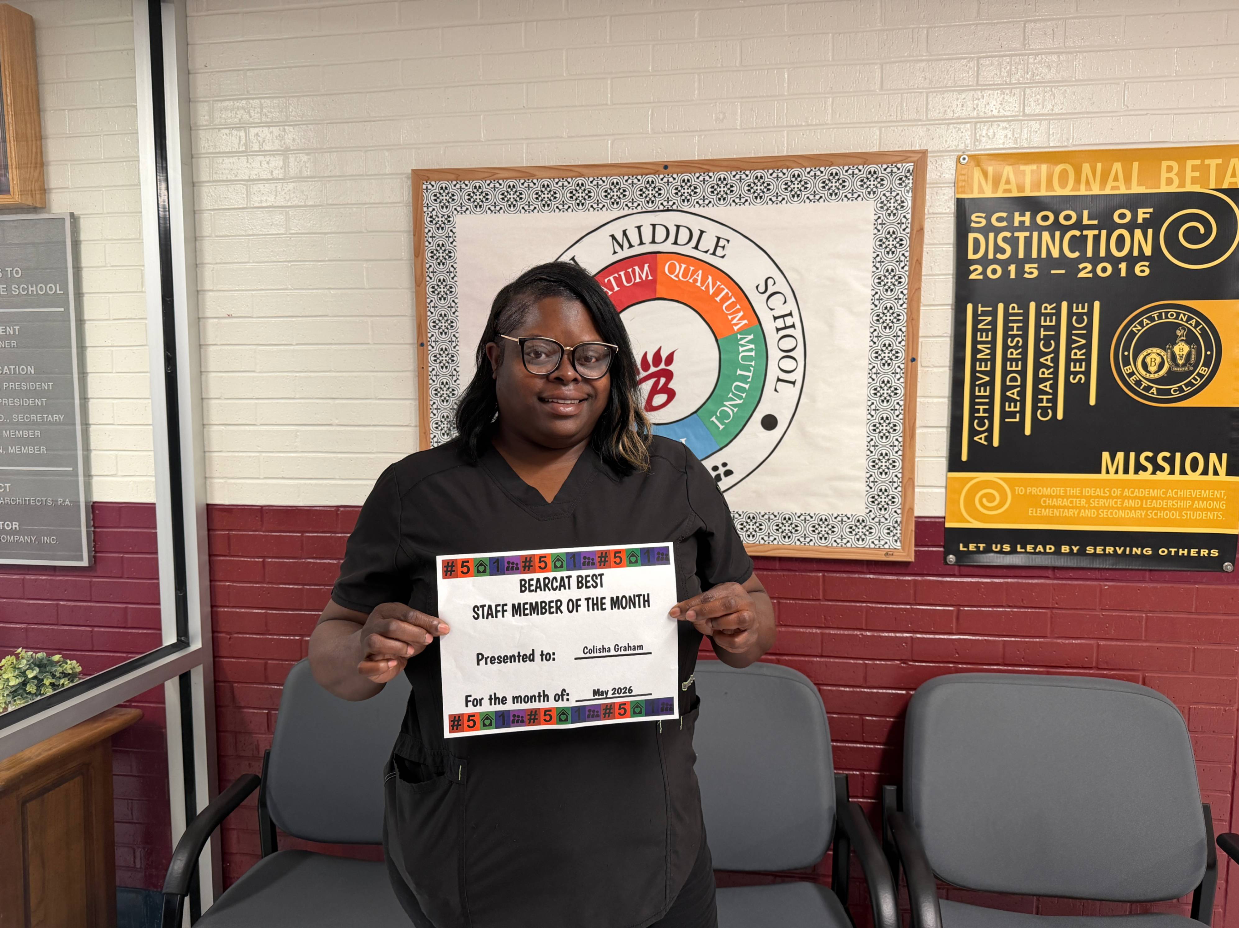 A person stands indoors in front of a white brick wall with school posters and banners, including a circular “Middle School” sign and a black-and-gold “National Beta School of Distinction 2015–2016” poster. The person is holding a certificate that reads “Bearcat Best Staff Member of the Month,” with the name “Colisha Graham” and “May 2026” visible. Several gray chairs line the wall behind the person.
