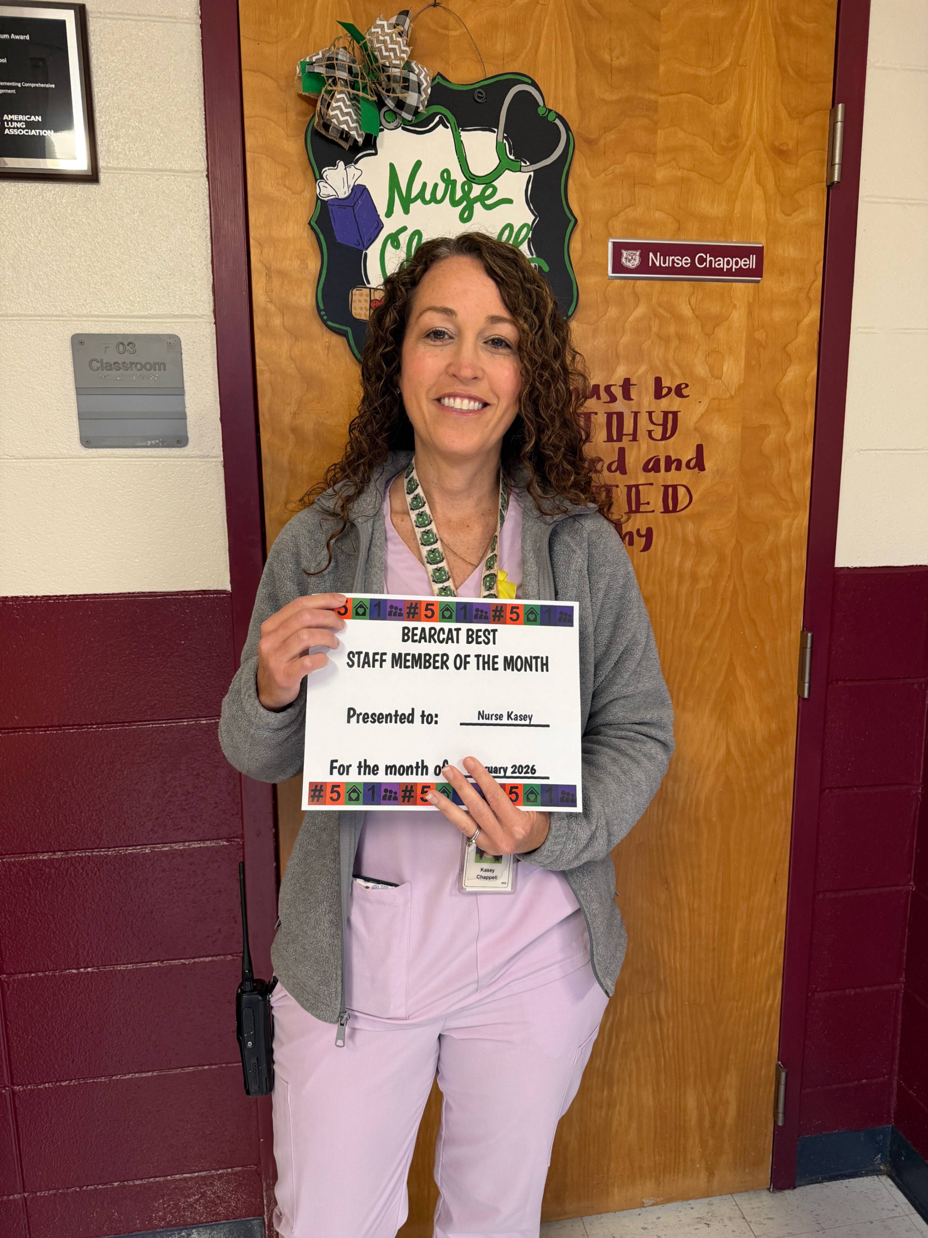 A woman stands smiling in a school hallway beside a wooden door labeled “Nurse Chappell.” She is holding a certificate recognizing her as the “Bearcat Best Staff Member of the Month,” presented to Nurse Kasey for January 2024. She is wearing light purple pants, a gray jacket, and a lanyard with an ID badge. The door behind her has a decorative medical‑themed sign with her name, and the wall next to it displays a plaque and a room sign labeled “703 Classroom.”