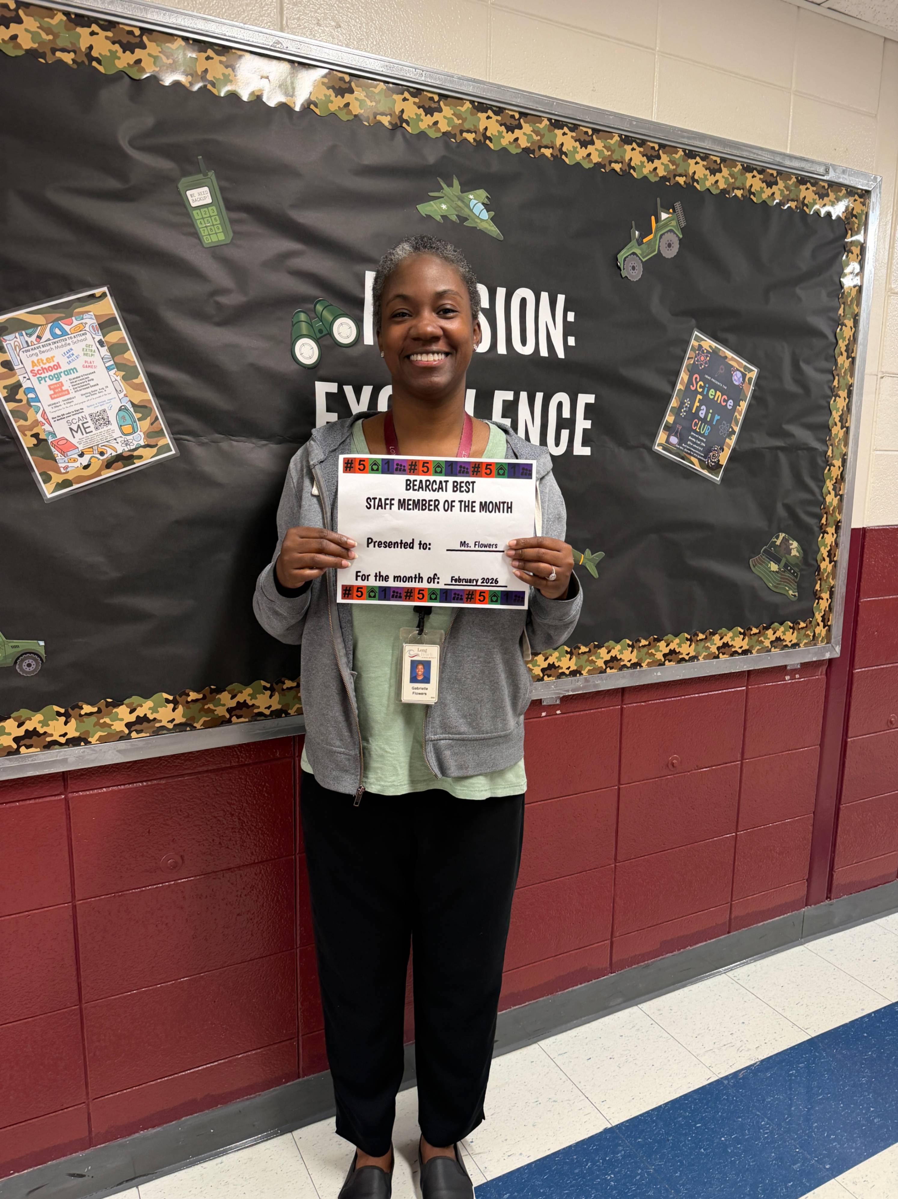    A woman stands smiling in front of a bulletin board decorated with a military theme, including camouflage borders and images of tanks, airplanes, and communication equipment. Large letters on the board read “Mission: Excellence.” She is holding a certificate that recognizes her as the “Bearcat Best Staff Member of the Month” for September, presented to Ms. Flowers.