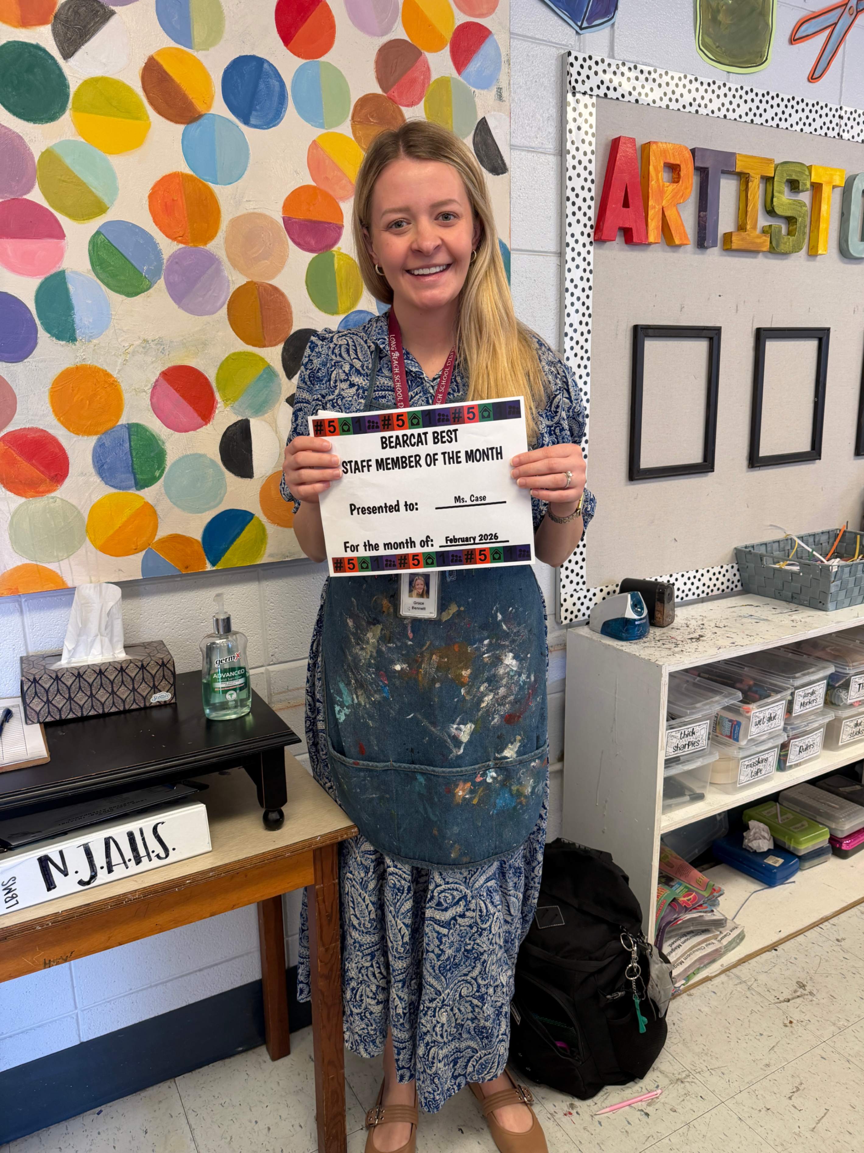A woman stands smiling in a colorful art classroom, holding a certificate. She is wearing a patterned dress and a paint‑stained apron. Behind her, the wall is decorated with multicolored circles and large letters spelling “ARTIST.” Art supplies and storage bins are visible around the room. The certificate reads “Berry Best Staff Member of the Month, Presented to Mrs. Case, February 2023.”